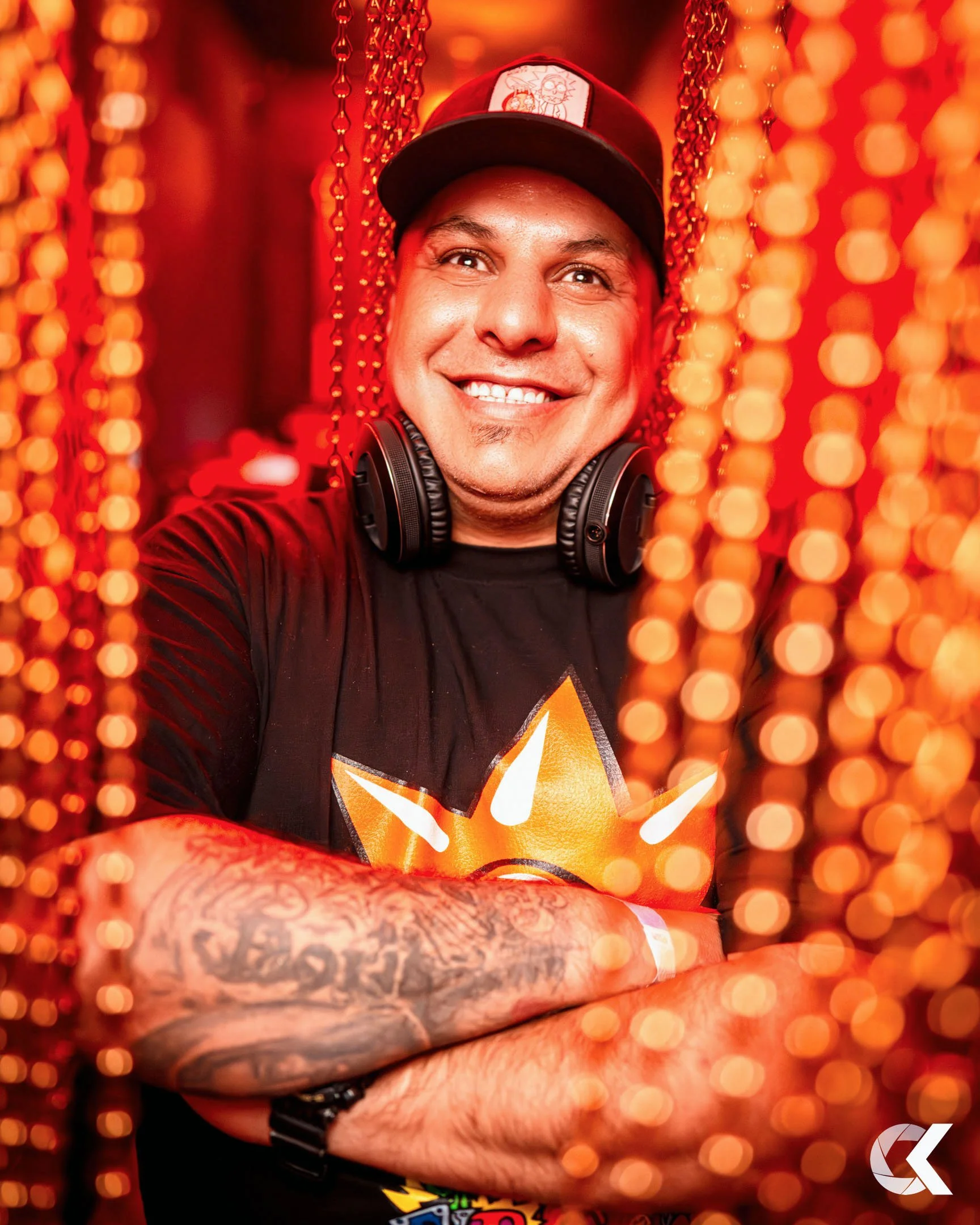 Man smiling, wearing a black T-shirt with a star logo, a black cap, and headphones around his neck, surrounded by orange beads and bokeh lights.