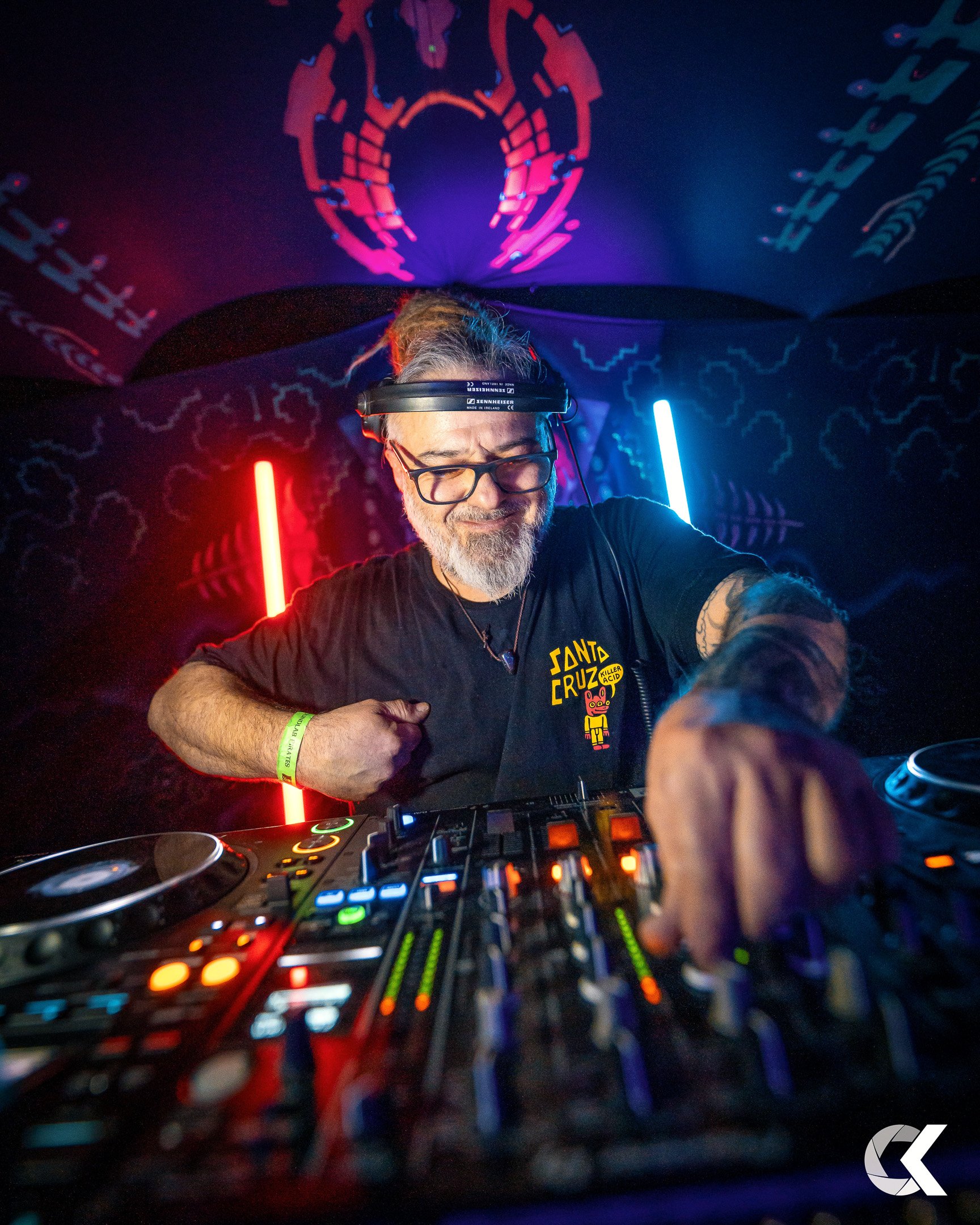 A DJ with glasses and a beard, wearing a black T-shirt with a colorful logo, is mixing music at a nightclub with vibrant red and blue lighting and psychedelic wall art in the background.