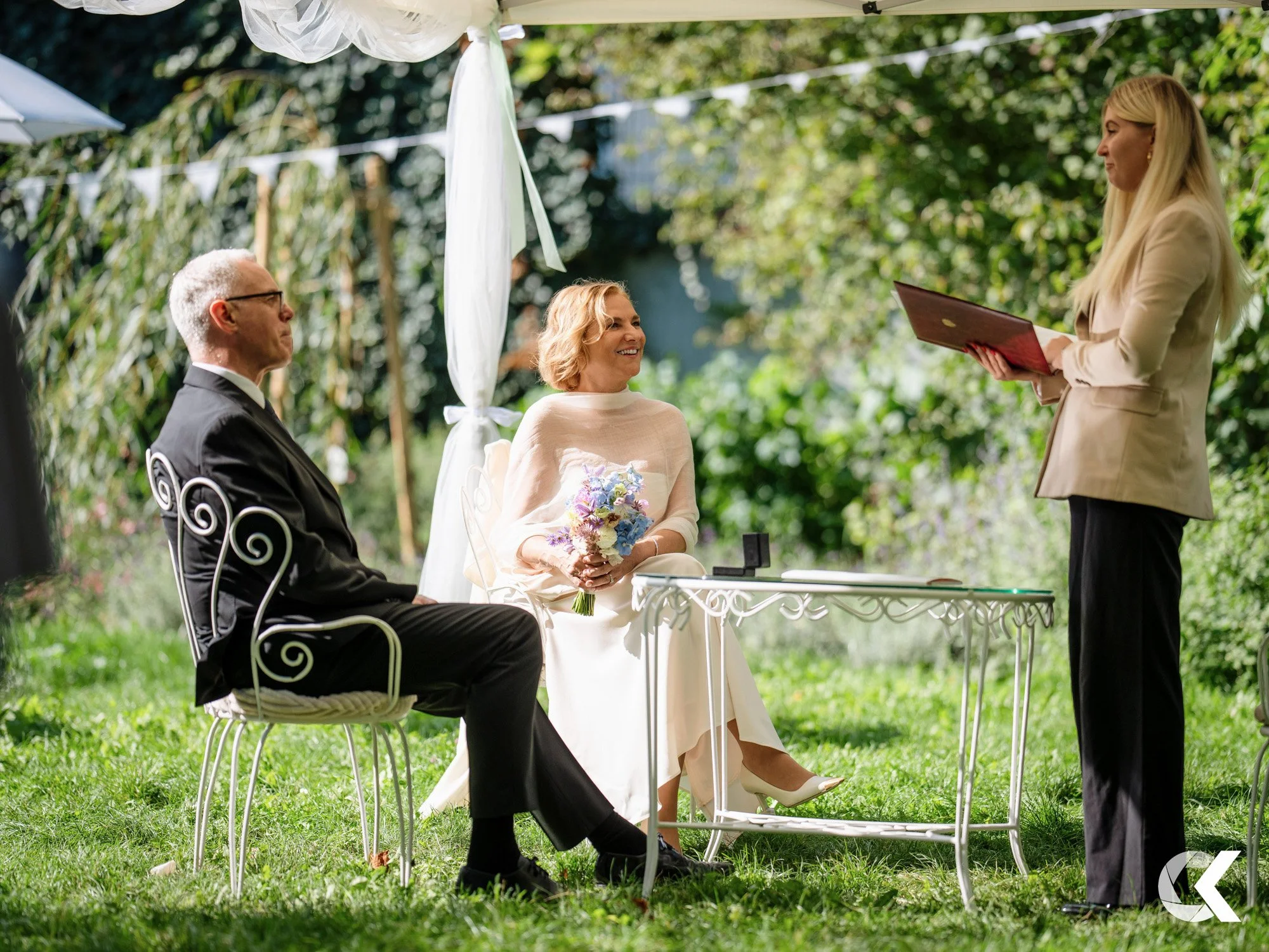 A woman in a white dress with a bouquet of flowers sits at an outdoor wedding ceremony under a white canopy, facing a woman in a beige blazer holding a book. A man in a black suit is seated next to her. The background is green with trees and sunlight