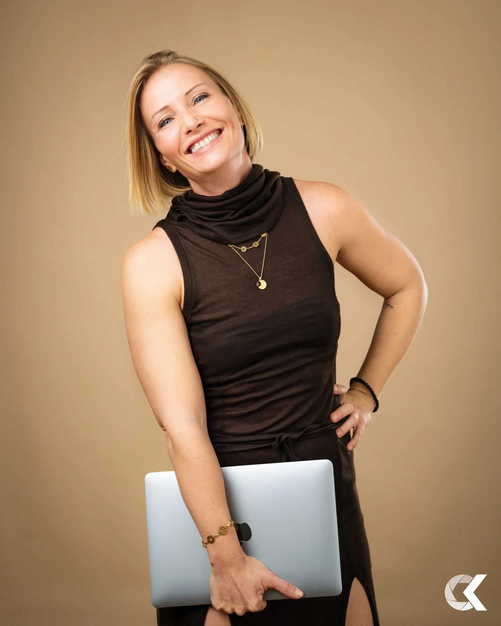 Smiling woman in black sleeveless top holding a laptop against beige background.