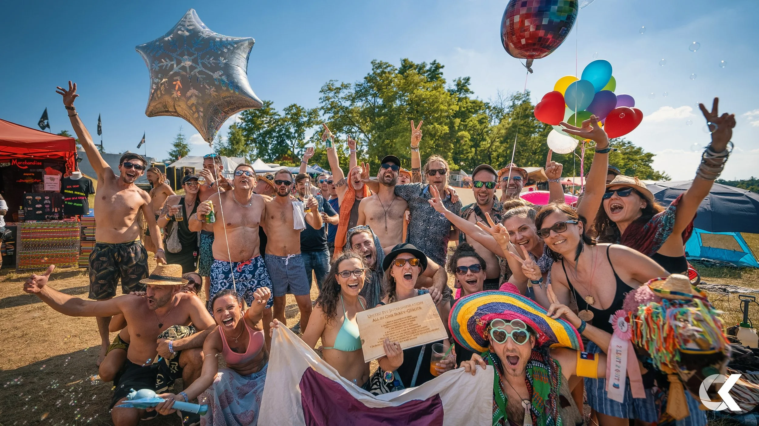 Group of people celebrating outdoors during summer festival, some wearing swimsuits, holding balloons, flags, and drinks, with tents and trees in background.