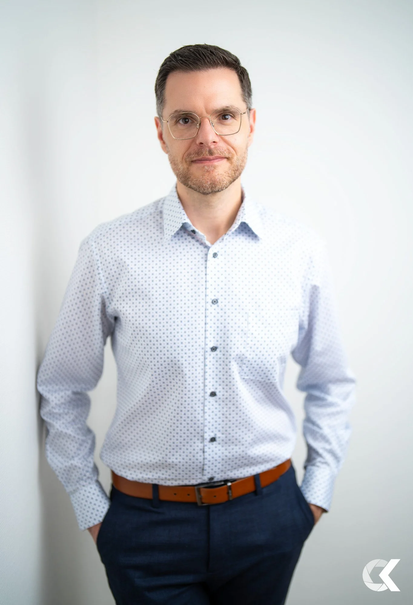 A man with dark hair and glasses wearing a light blue patterned shirt and dark pants, standing against a white wall.