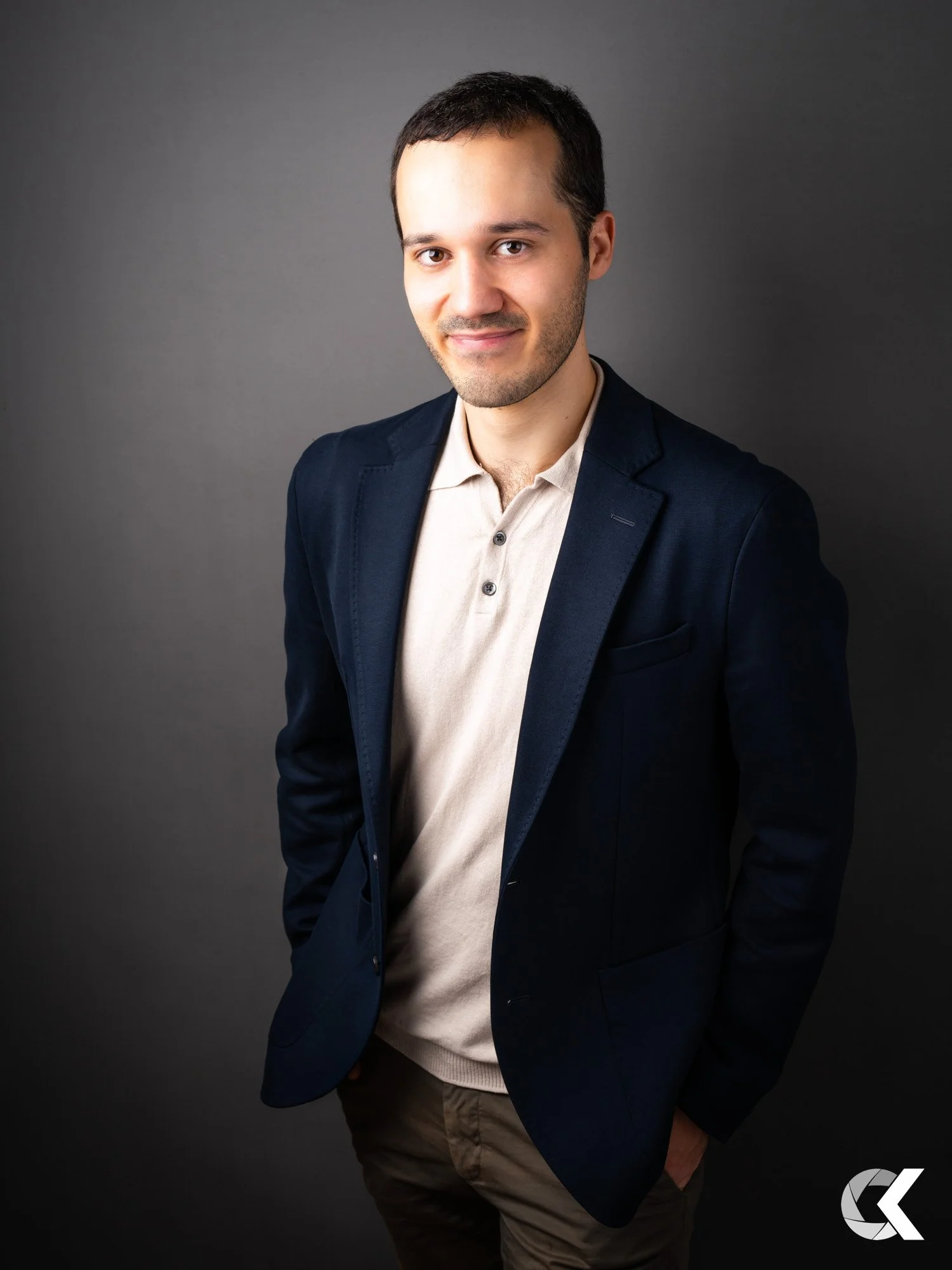 Portrait of a young man with dark hair, wearing a beige polo shirt and navy blazer, standing with his hands in his pockets against a gray background.