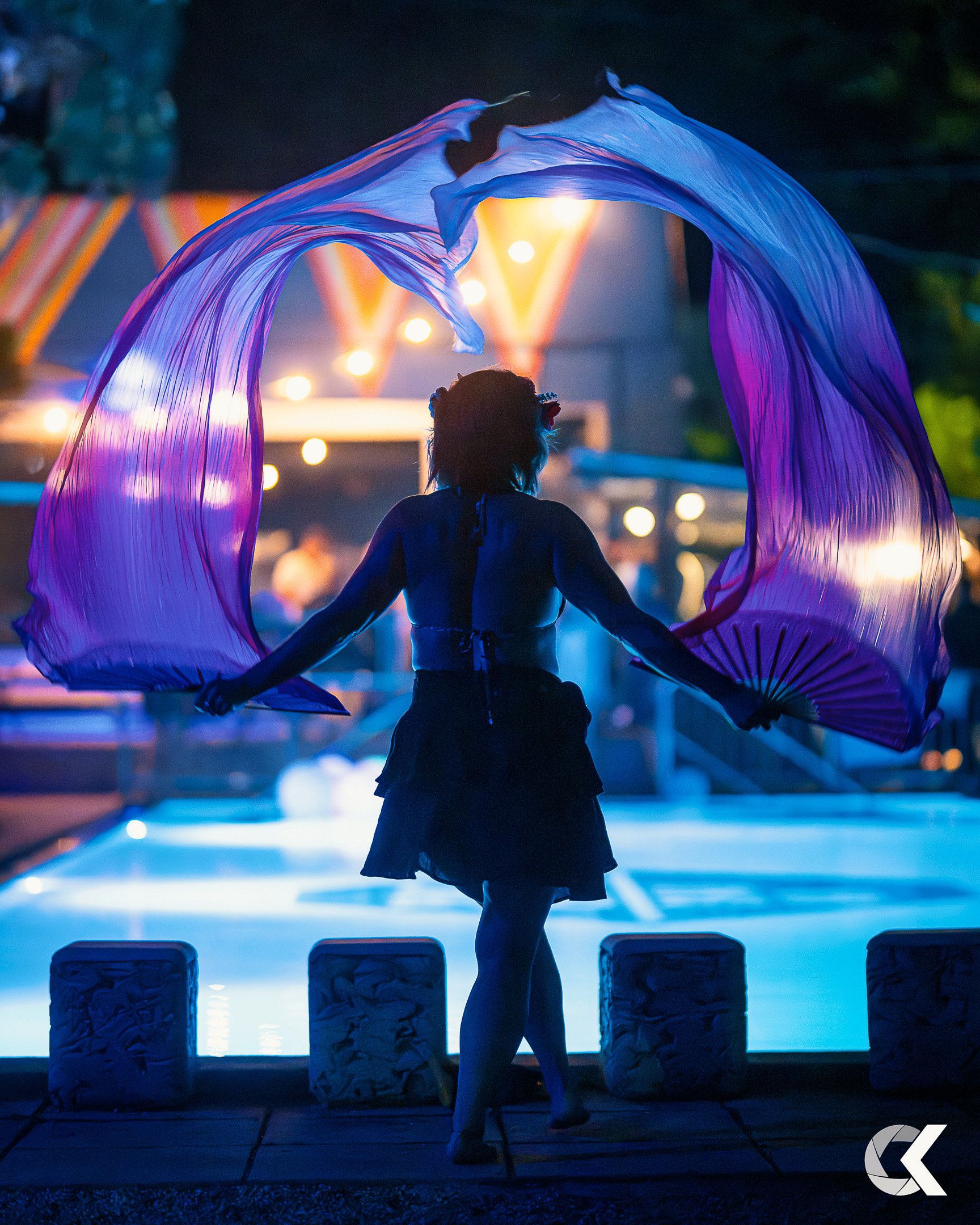 Silhouette of a person holding purple and pink flowy fabric, standing near a pool at night illuminated by colorful lights.
