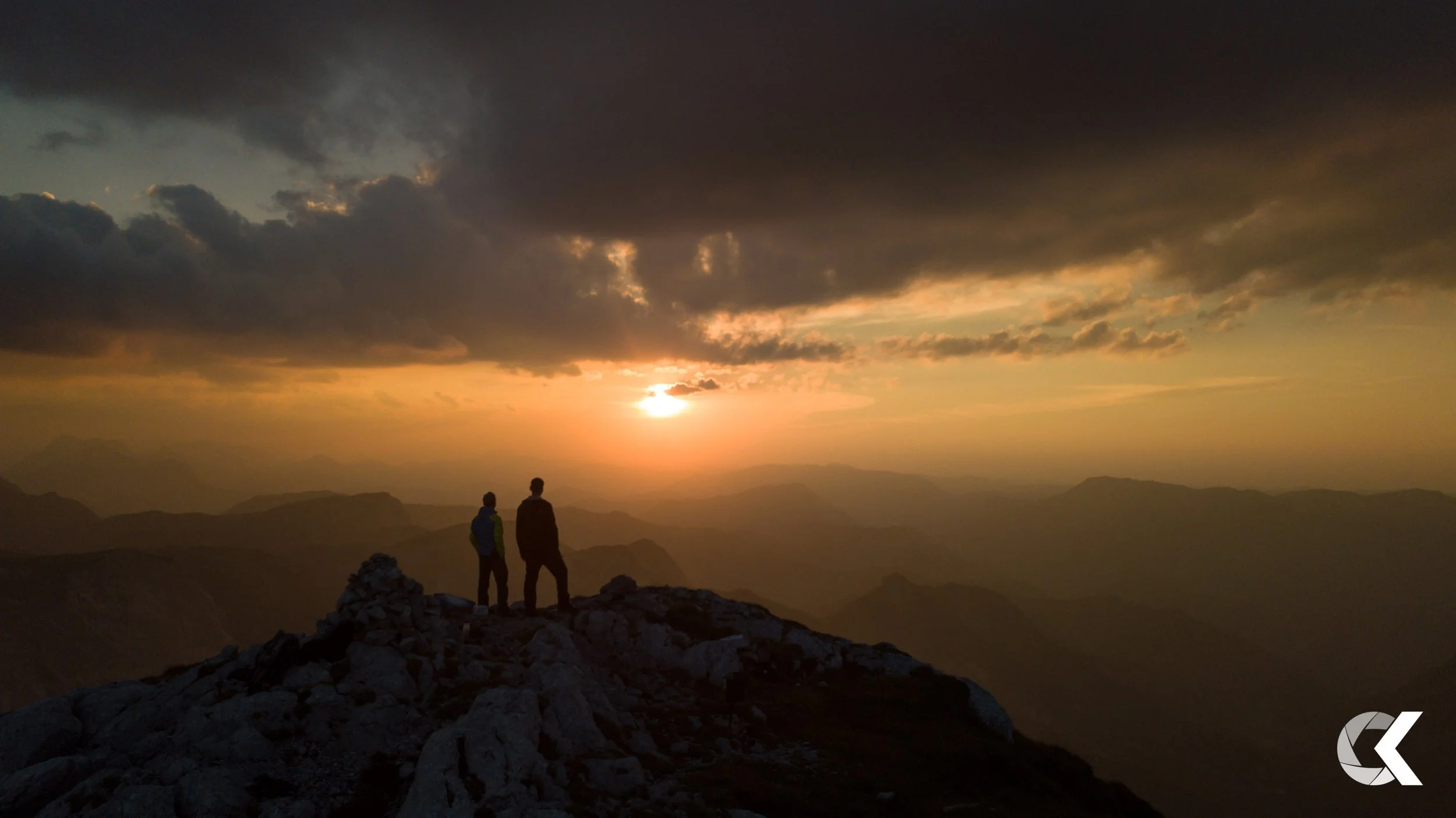 Two hikers stand on a rocky mountain peak, watching a sunset over distant mountain ranges with dark clouds overhead.