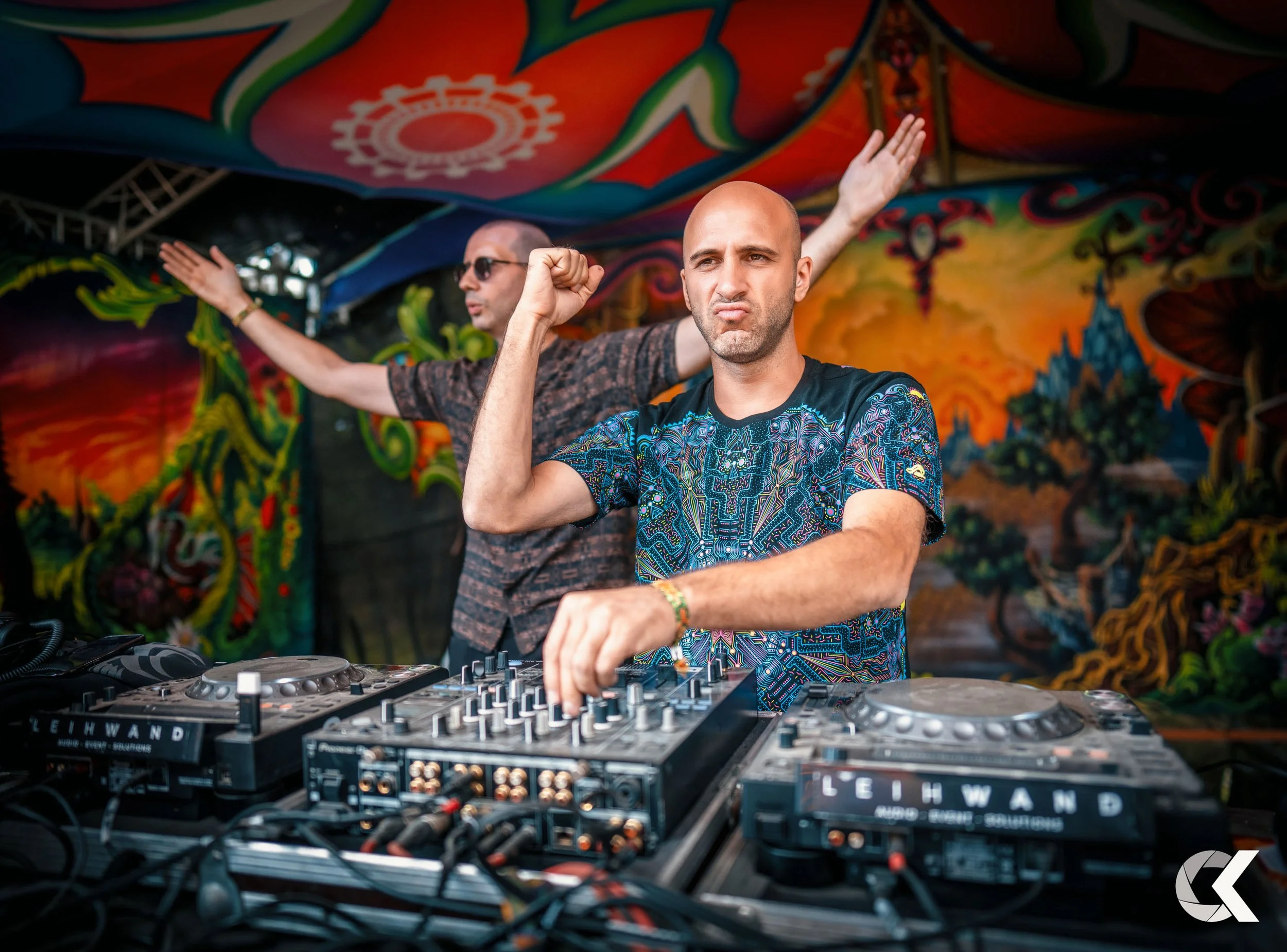 Two men performing as DJs at a colorful, psychedelic festival. The man in the foreground, with a shaved head, wears a vibrant patterned shirt and is making a fist pump gesture. The man in the background, with sunglasses, extends his arms. The scene i