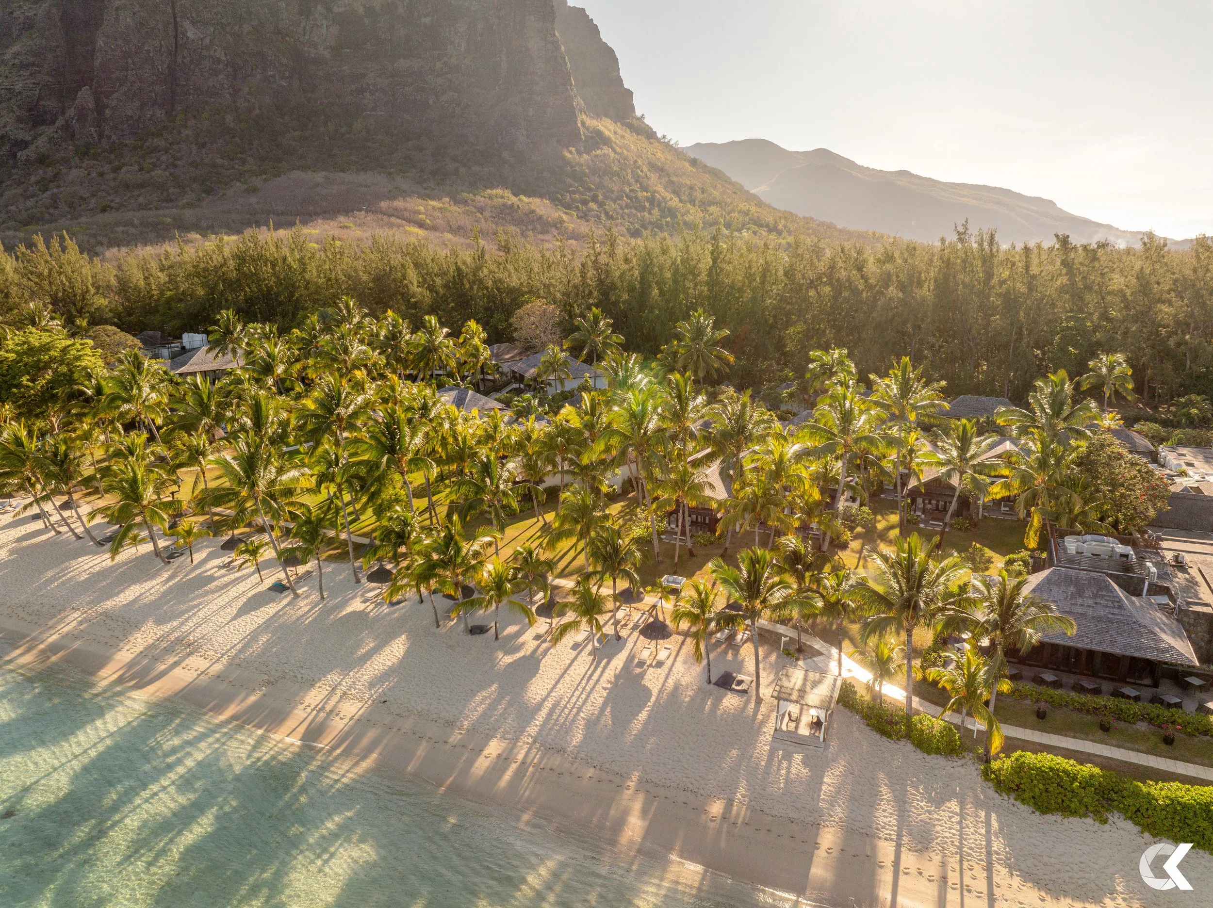 Aerial view of a tropical resort with palm trees, white sandy beach, and clear ocean water, with mountains in the background during sunset.