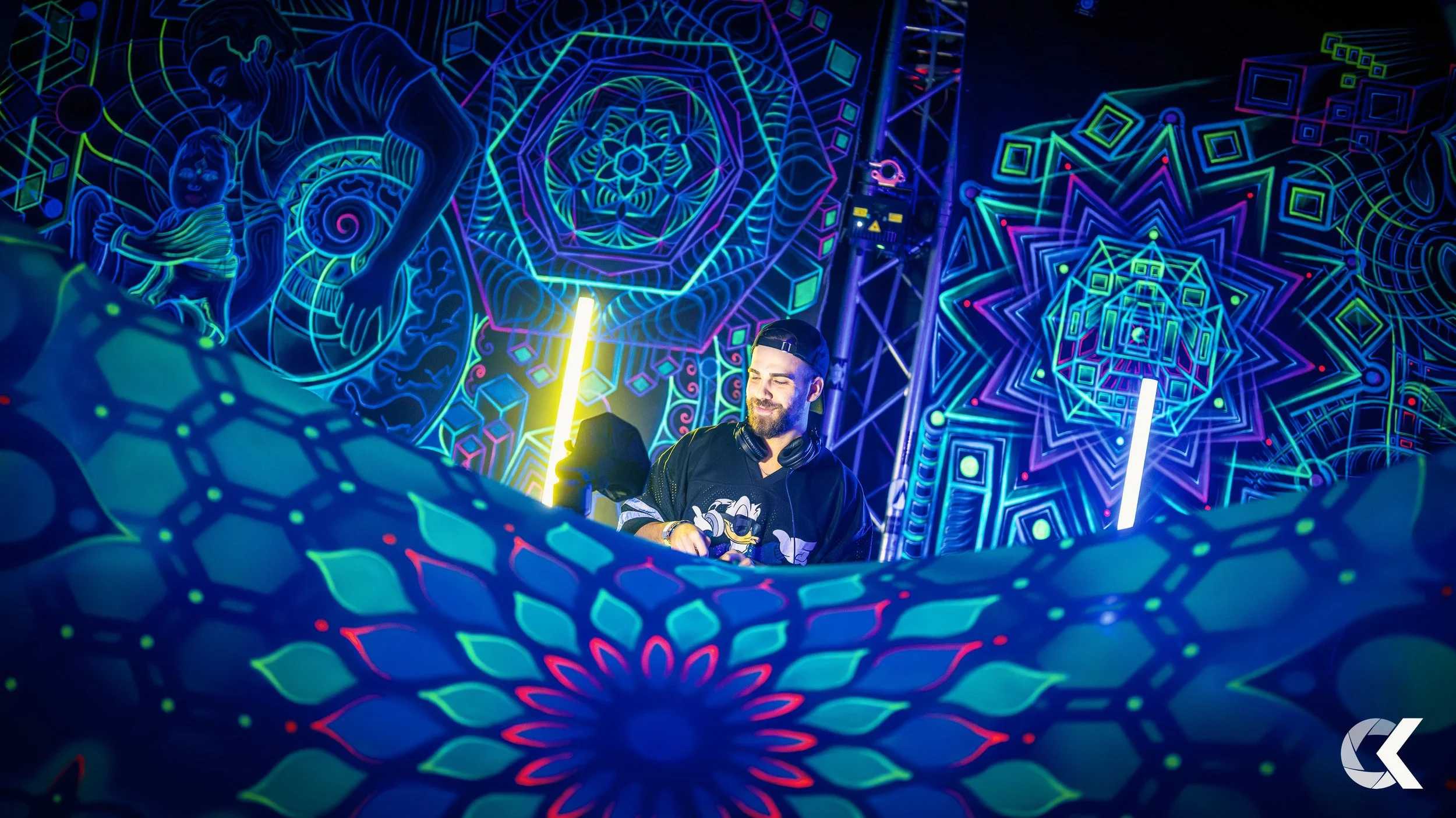 A DJ with a beard, wearing a black cap backwards and headphones around his neck, is performing at a neon-lit event. The backdrop features colorful, intricate geometric and mandala designs illuminated by glow-in-the-dark and LED lights.