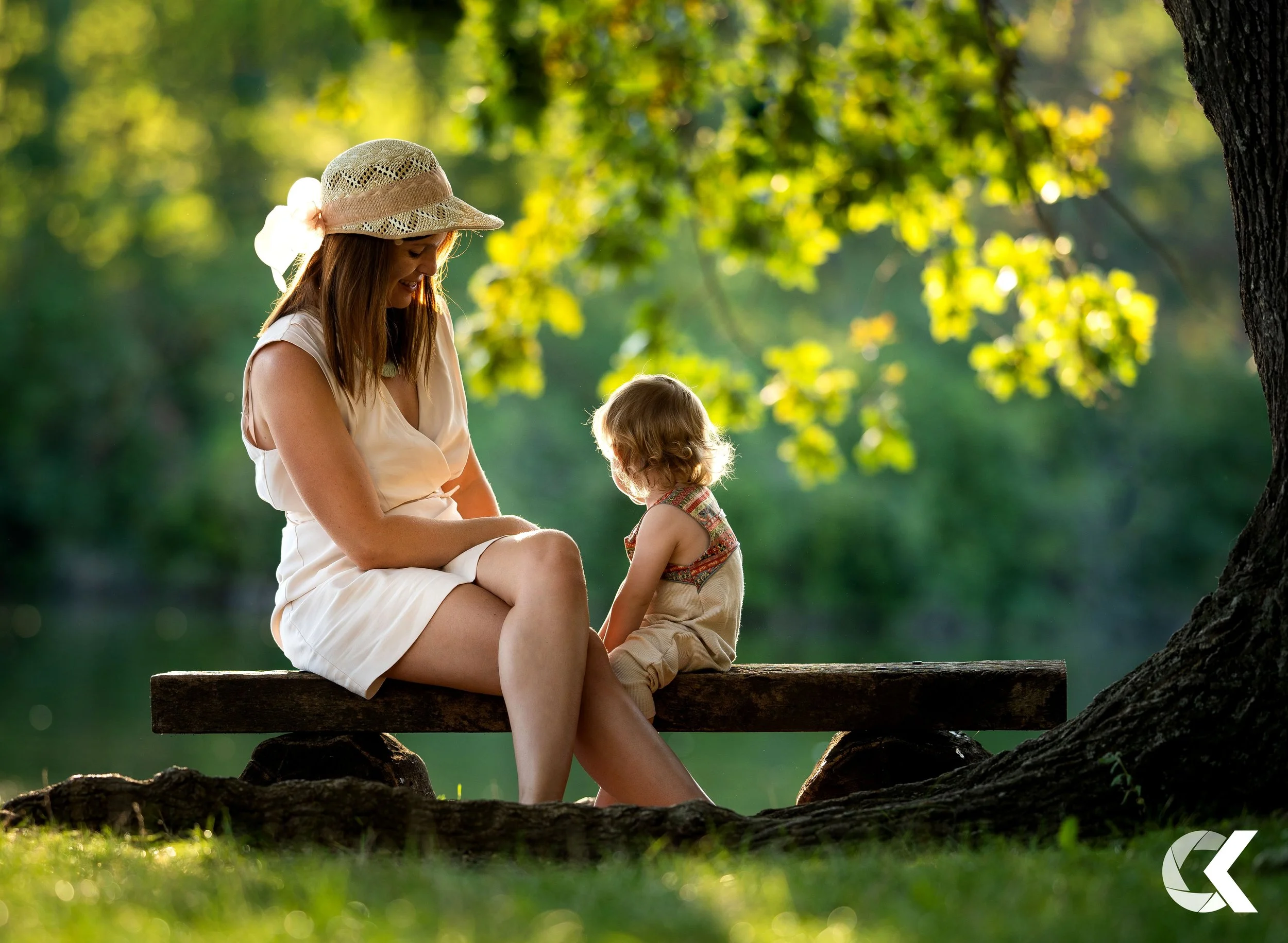 A woman and a young girl sitting on a wooden bench under a large tree, outdoors, with sunlight filtering through green leaves.