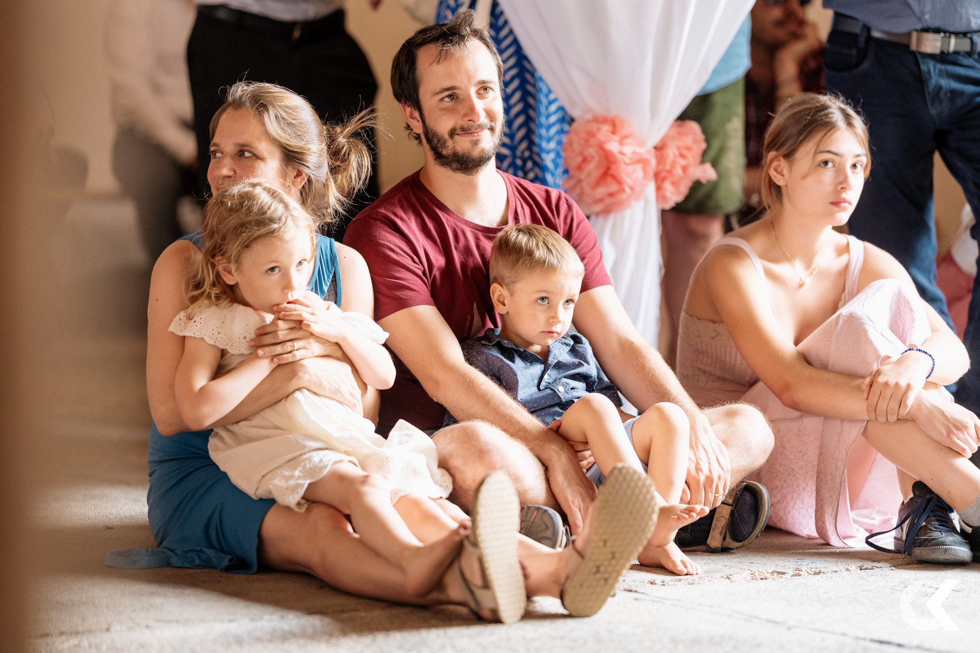 A group of people sitting on the floor, including a woman with a young girl in her lap, a man, a boy, and a young woman, at what appears to be a social gathering or event.