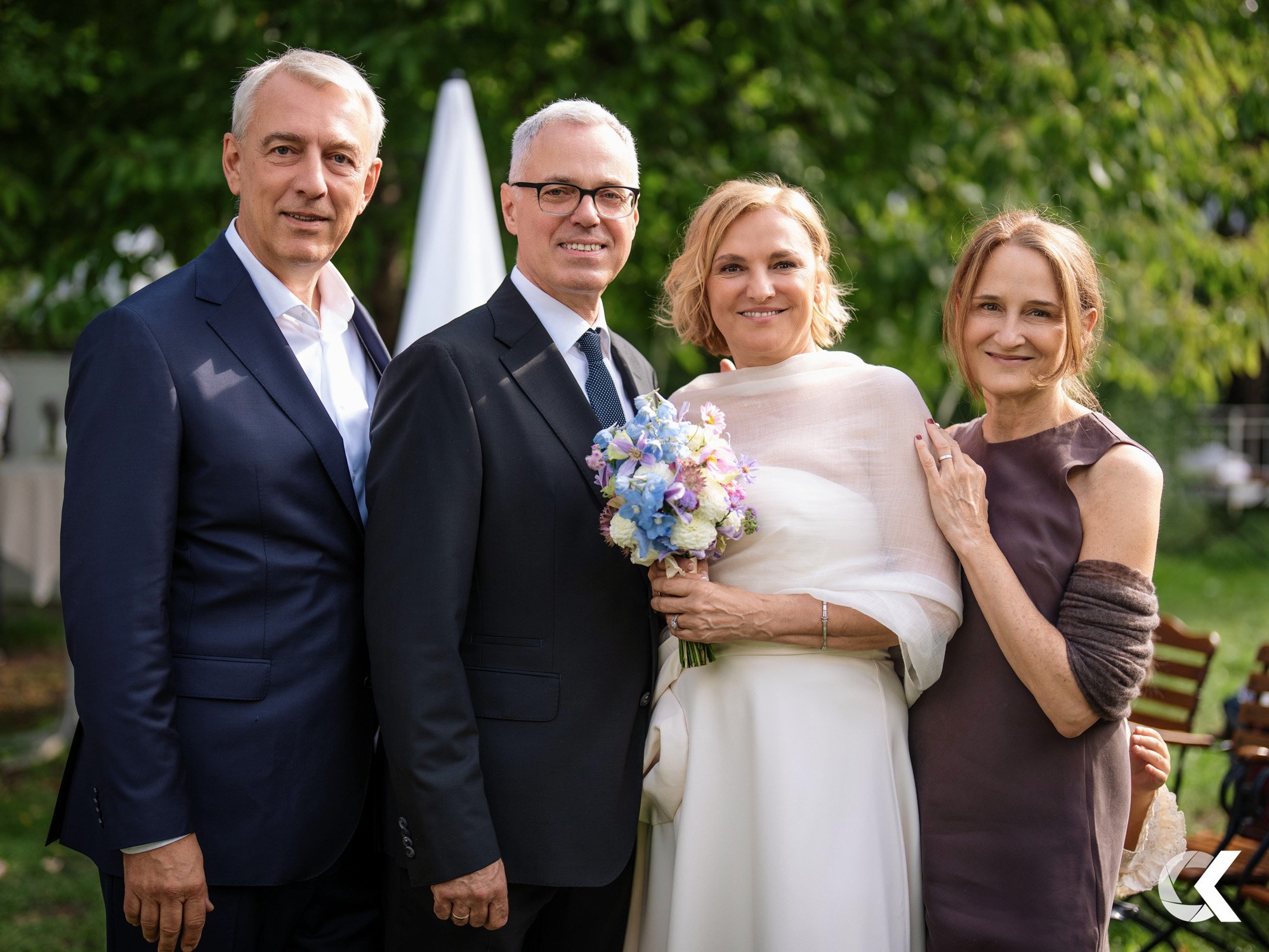 Four people smiling outdoors at a wedding, with trees and chairs in the background. Two men in suits, a woman in a white dress holding a bouquet, and another woman in a brown dress.