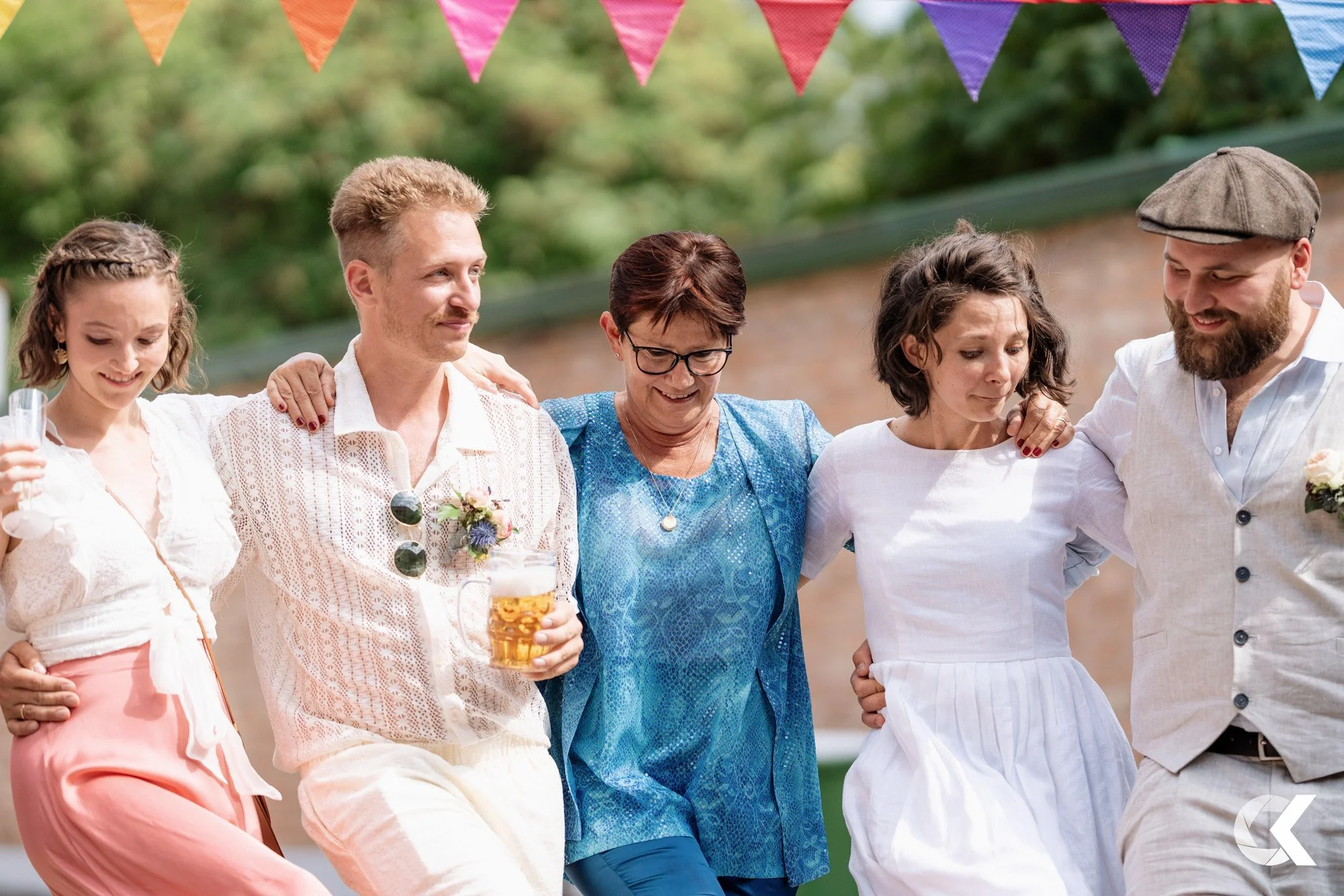 Six people are standing closely together outdoors with their arms around each other, at a social gathering or party. Colorful bunting hangs overhead, and they are dressed in casual summer clothing.