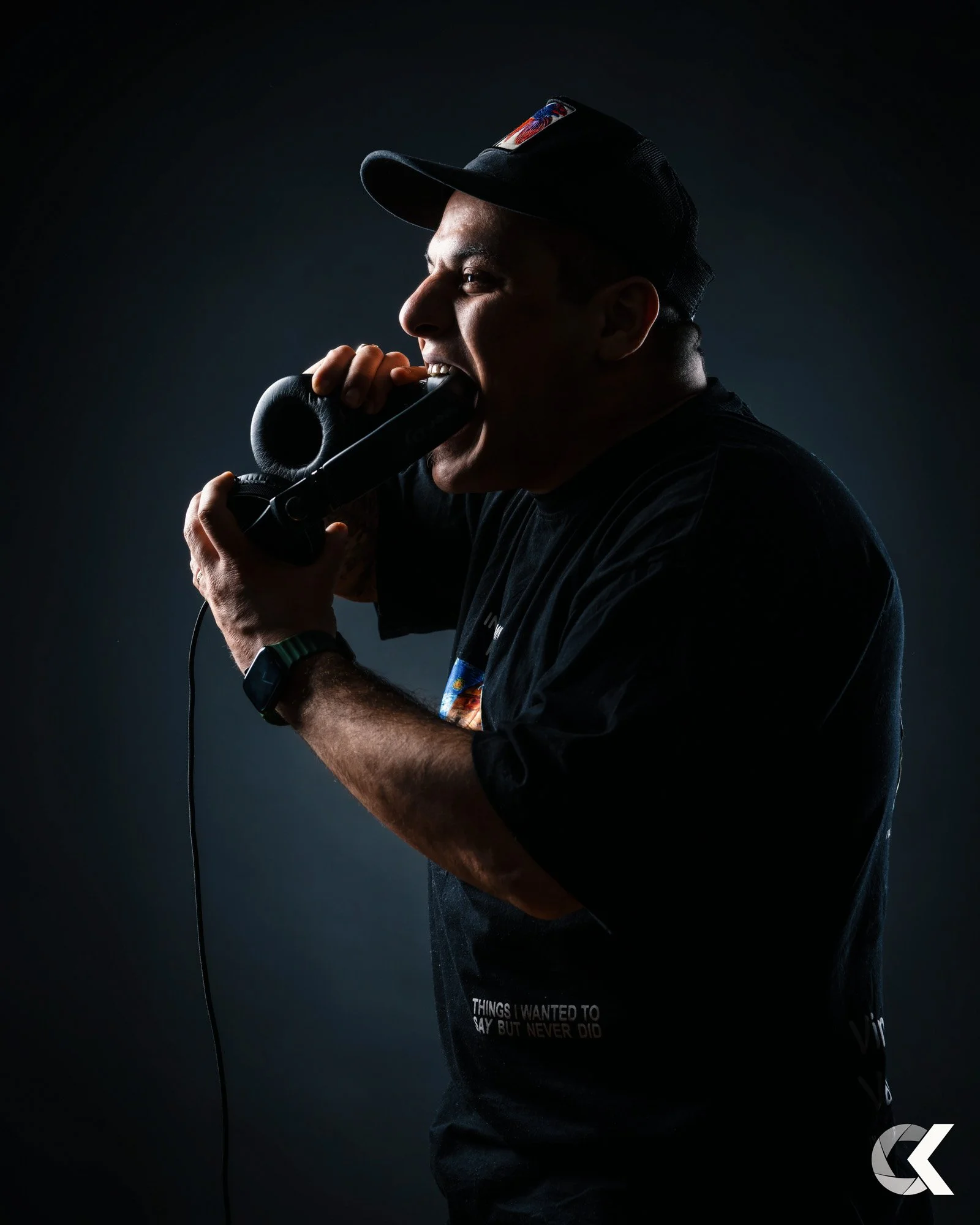 A man in a dark shirt and cap biting a telephone receiver, set against a dark background.