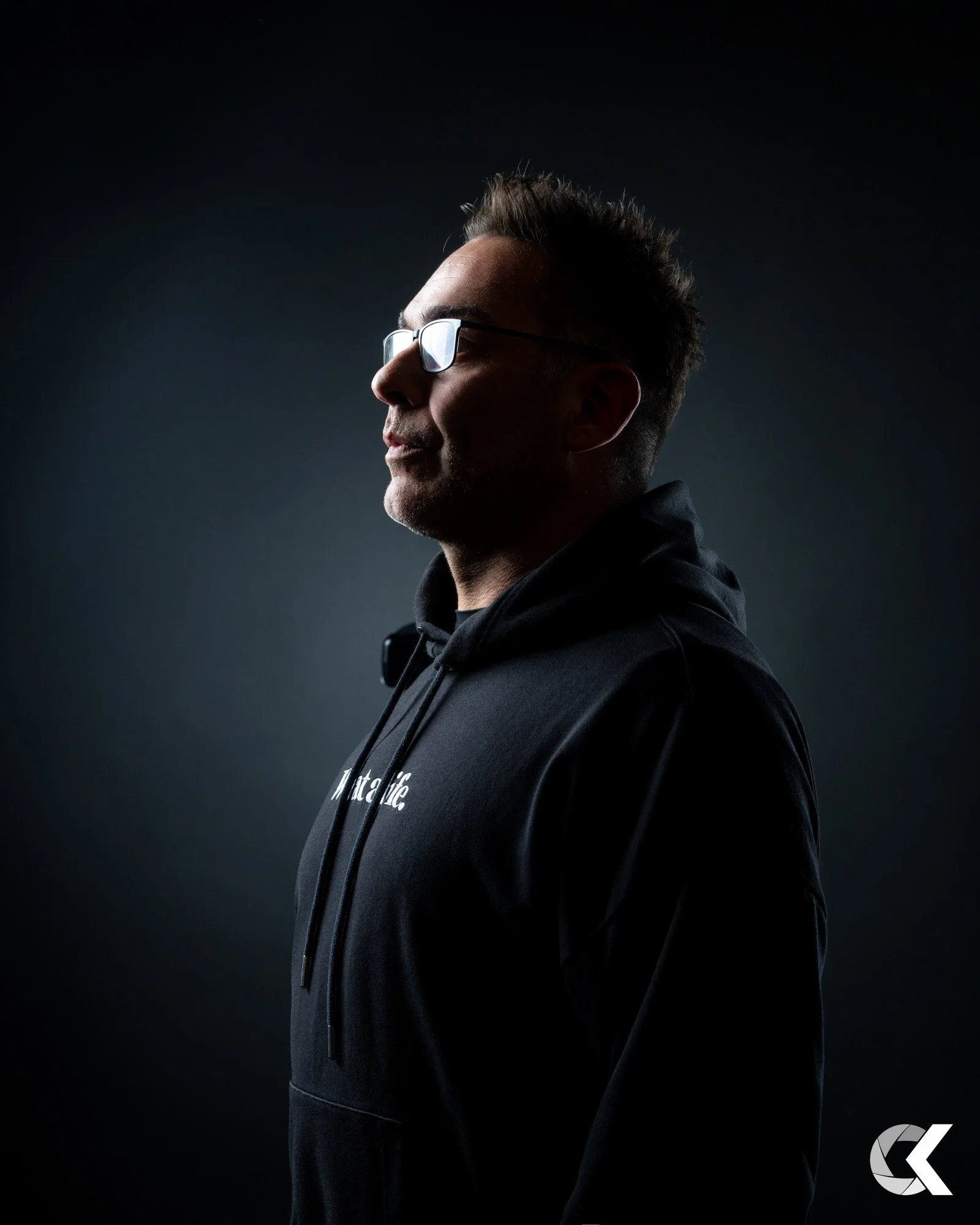 Side profile of a man wearing glasses and a black hoodie against a dark background.