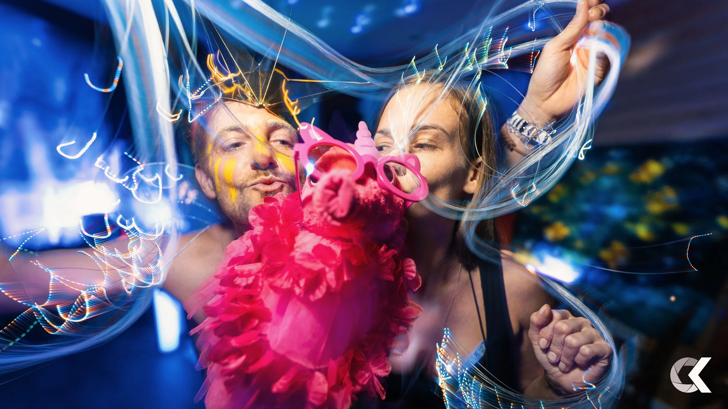 A man and woman taking a selfie at a party with colorful light trails, a pink unicorn headband, and a pink flower hair accessory.