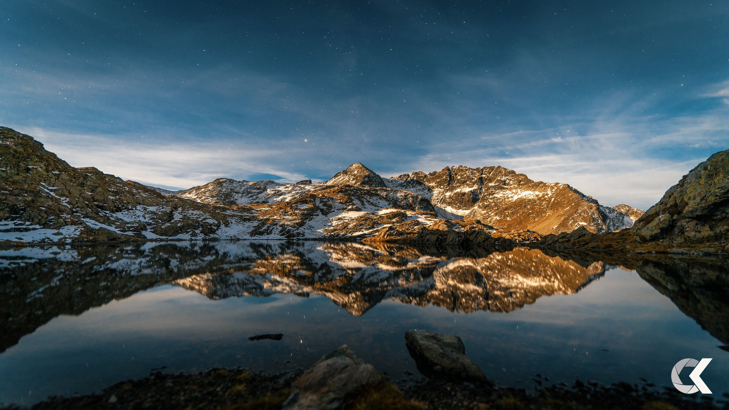 A mountain landscape at dusk with snow-capped peaks reflected in a calm lake, under a starry sky.