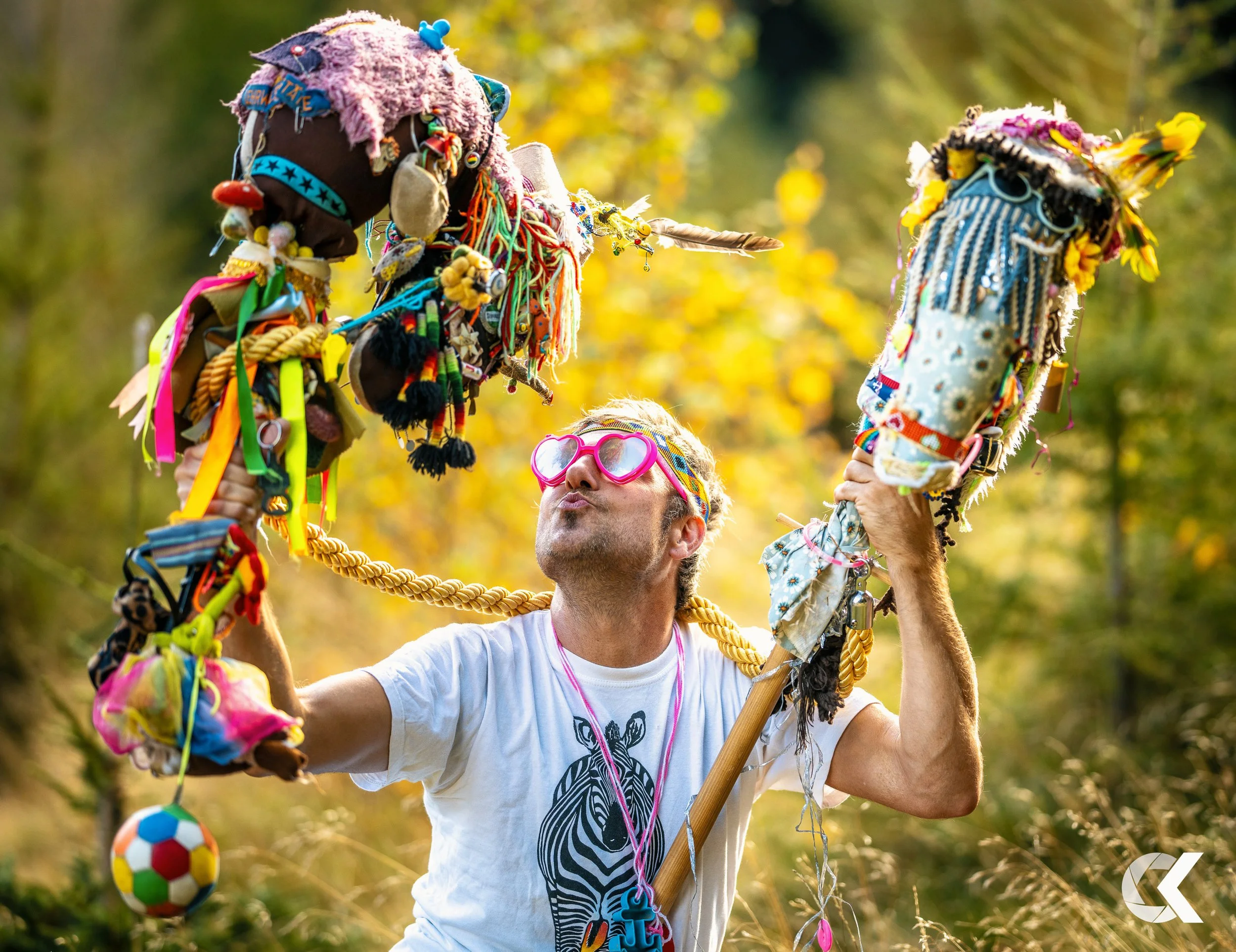 Man with pink sunglasses holding two decorated sticks with colorful, handmade crafts and face puckered in a kissing pose outdoors with green trees and sunlight.