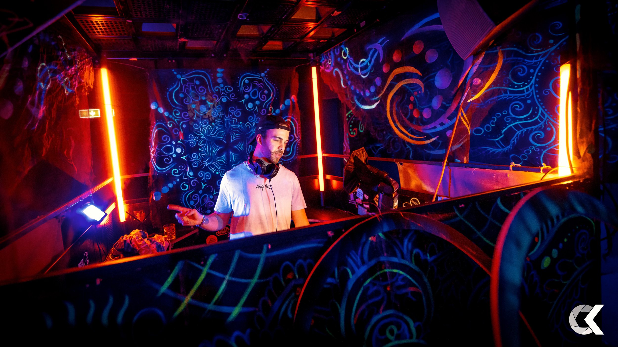 A DJ in a white t-shirt with headphones around his neck performing at a nightclub with neon and blacklight-lit graffiti-style artwork on the walls.