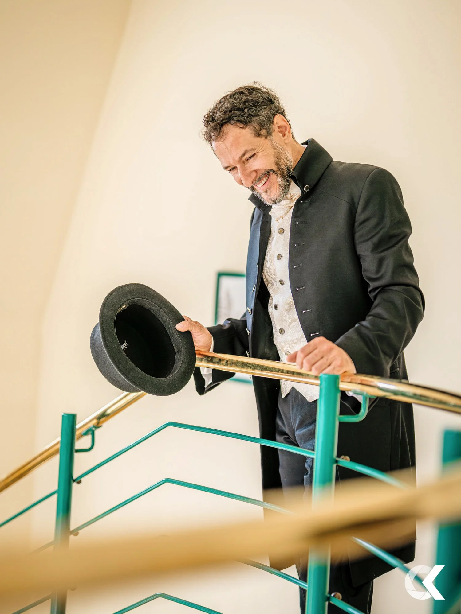 A man dressed in a historical costume, holding a black top hat and a long stick, smiling and looking downward, standing behind a green metal railing.