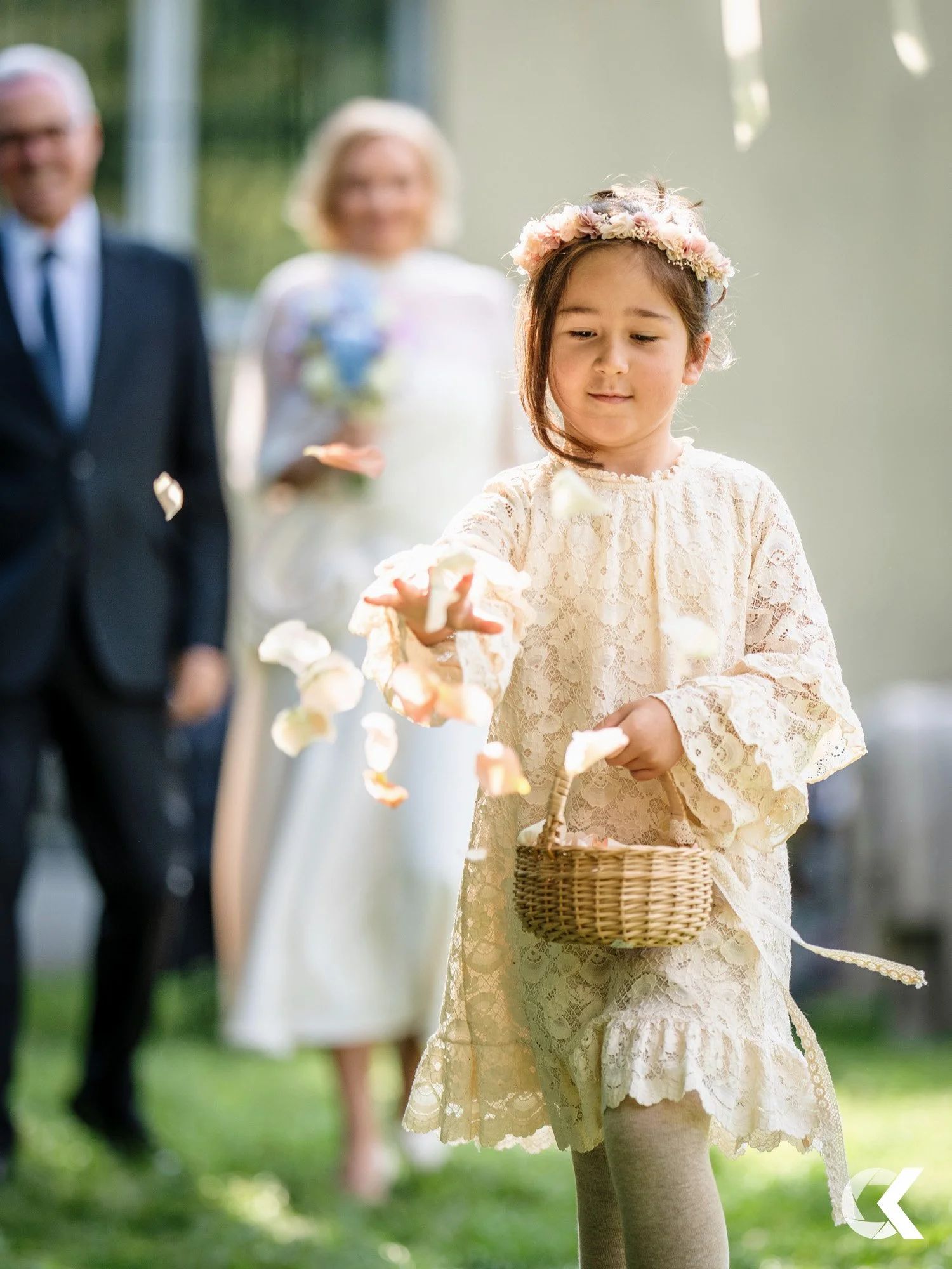 Young girl in lace dress and floral crown picking flower petals, holding a basket, at an outdoor event with adults in the background.