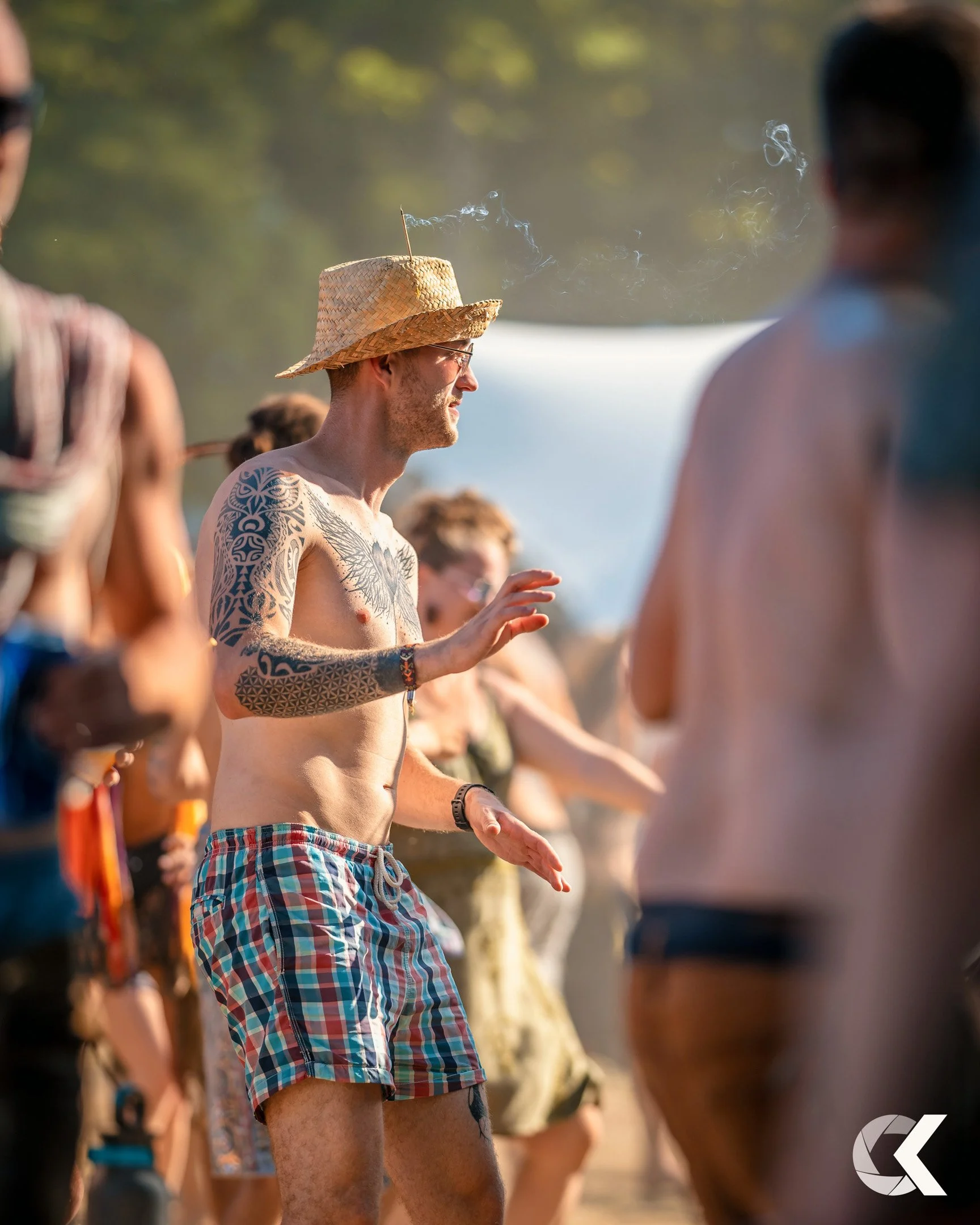 Group of people dancing outdoors in summer, with a man wearing a straw hat, colorful checkered shorts, and tattoos, surrounded by others in casual summer clothing.