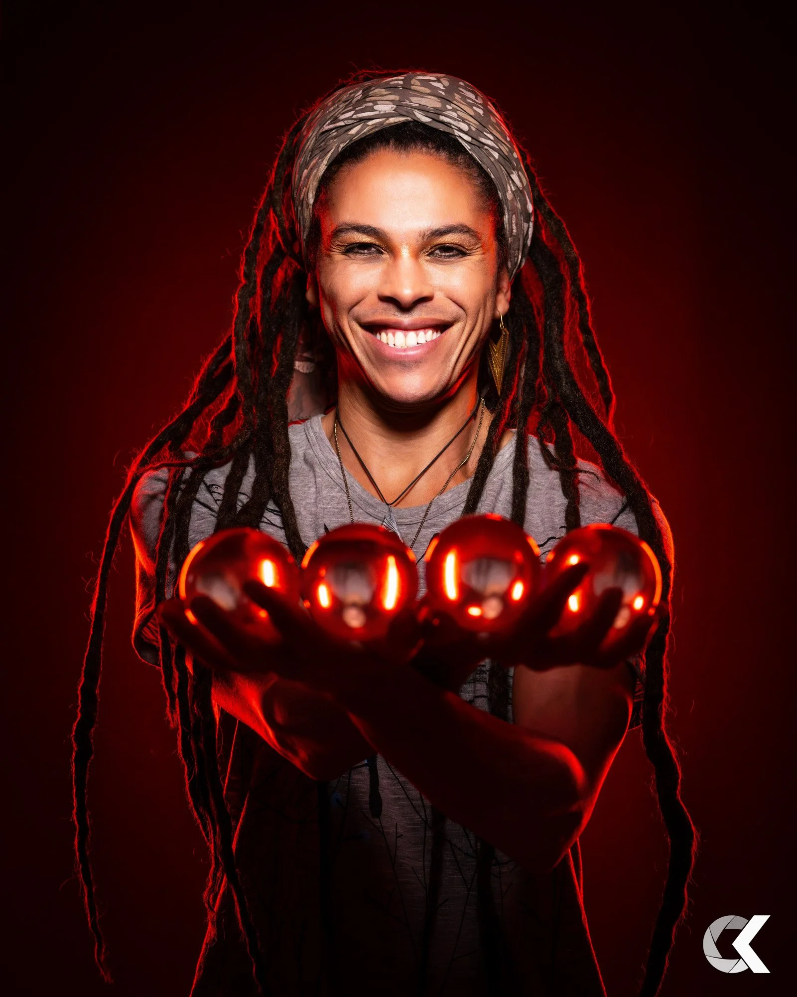 Person with dreadlocks holding red Christmas ornaments, smiling, wearing a headscarf and gray shirt, with red lighting in the background.