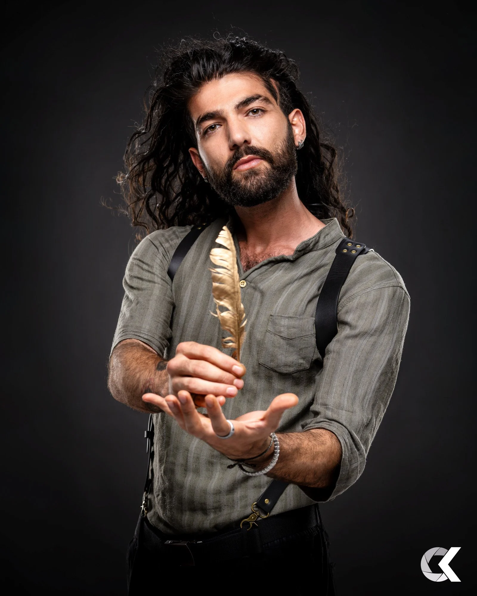 A man with long curly hair and a beard is holding a gold-colored feather in his hand, looking at the camera, against a dark background.