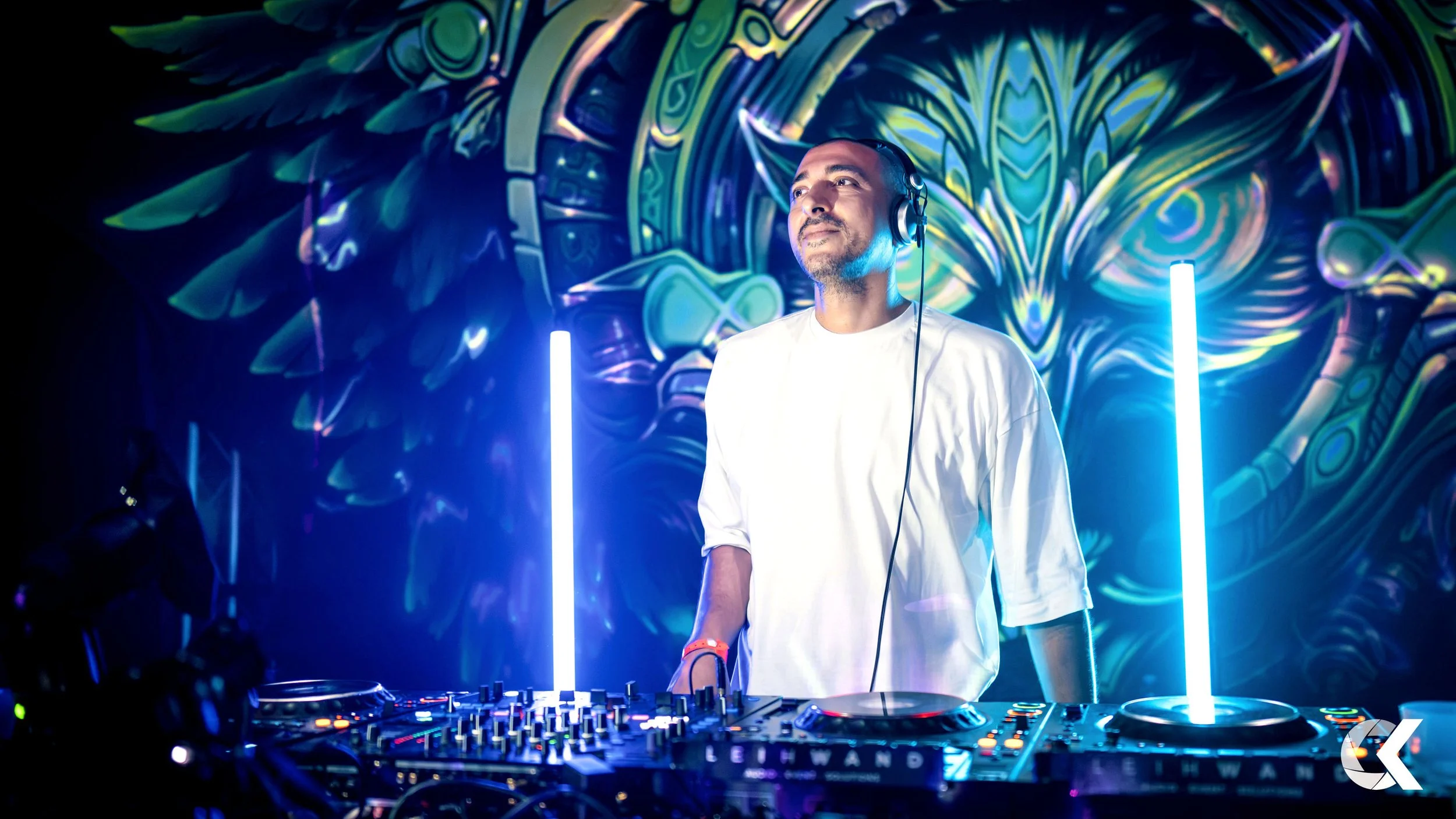 A DJ wearing a white shirt and headphones stands behind a DJ console with vivid, colorful, and abstract graffiti-style artwork in the background, illuminated by blue and purple lighting.