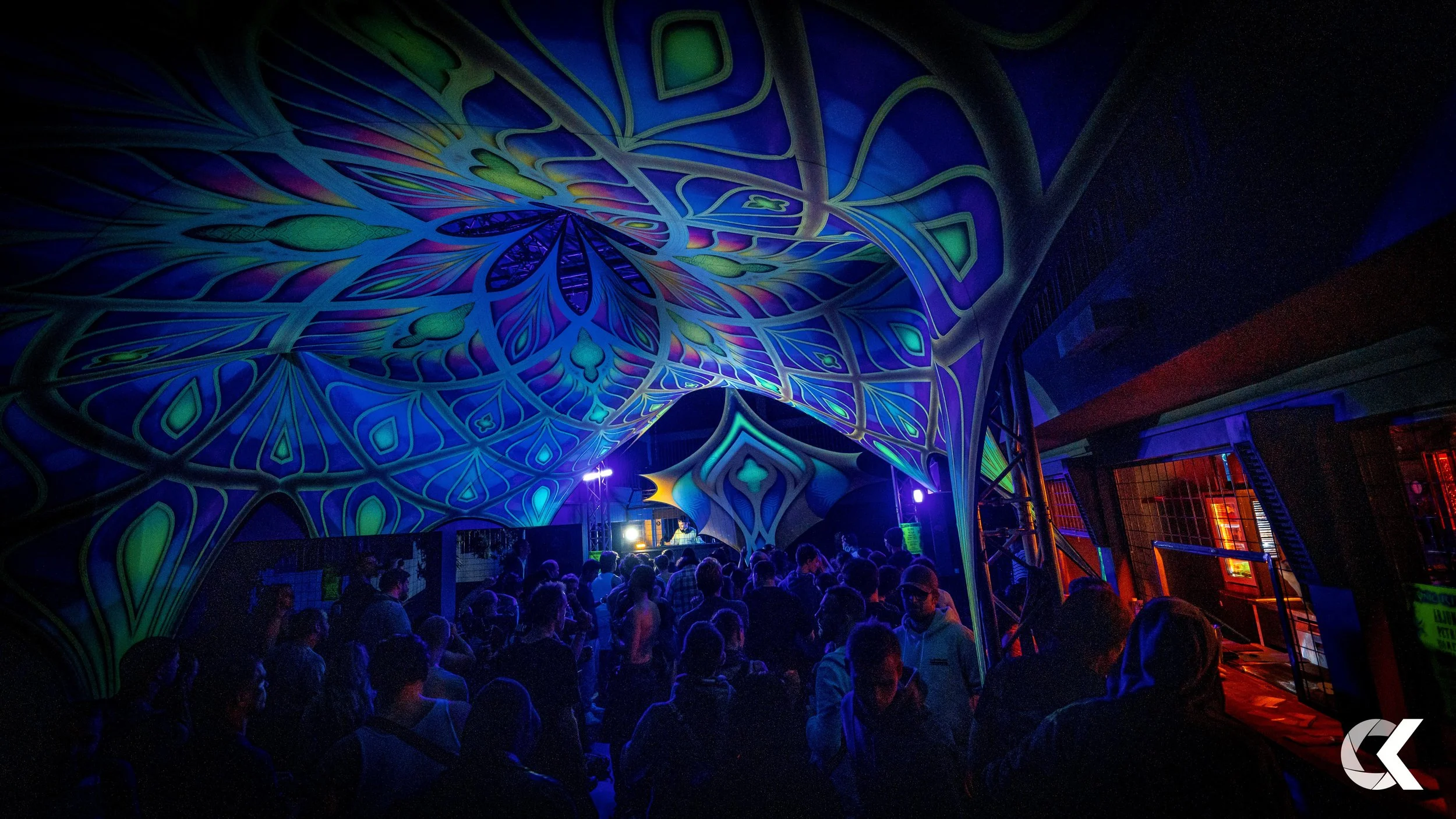 Crowd of people at a music festival or nightclub, under a large colorful geometric and abstract draped fabric, illuminated by vibrant blue, green, purple, and yellow lights, with DJ booth in the background.
