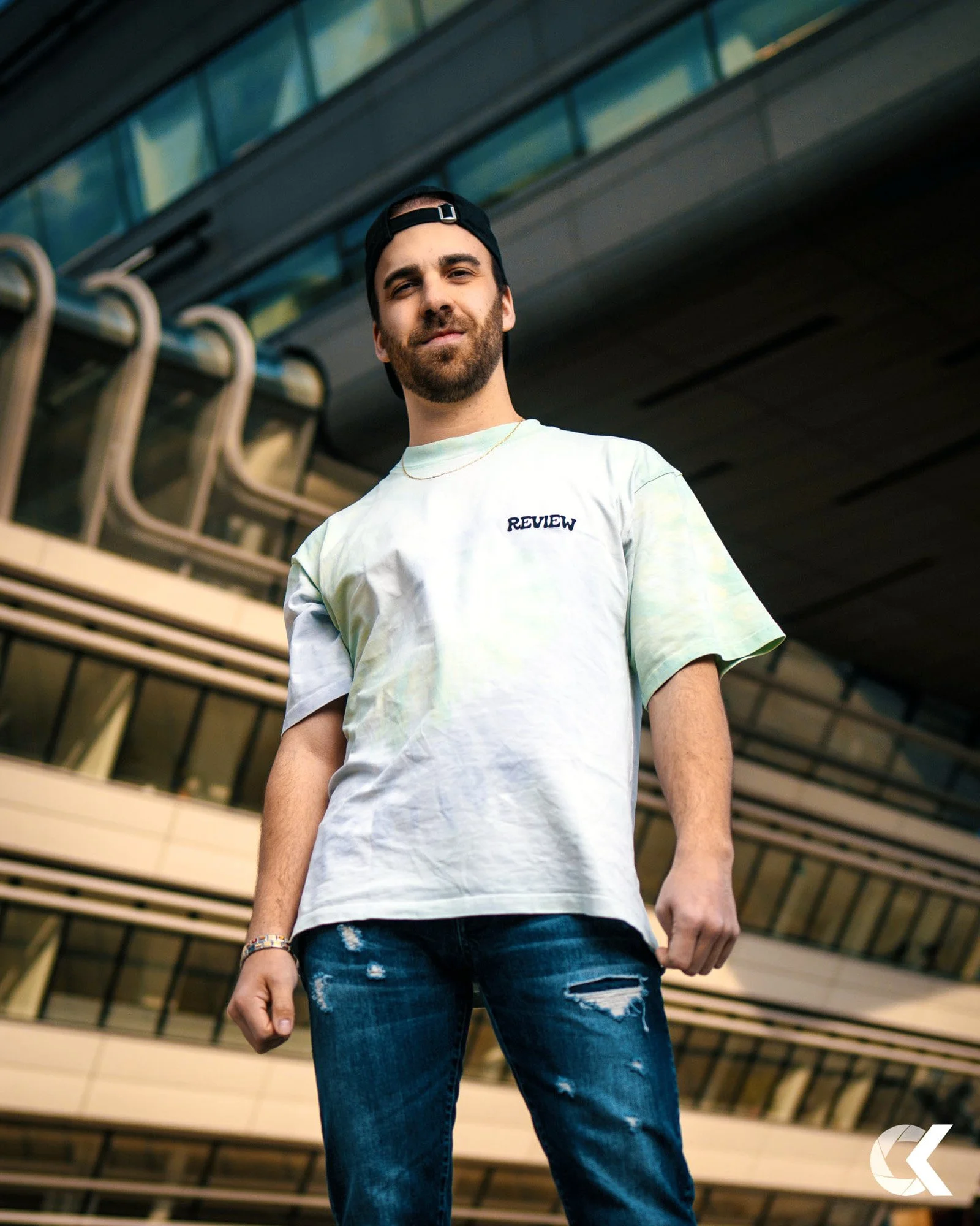 A young man with dark hair and beard wearing a black cap backwards, a white T-shirt with 'REVIEW' written on it, and ripped jeans, standing outdoors in front of a modern building with balconies.