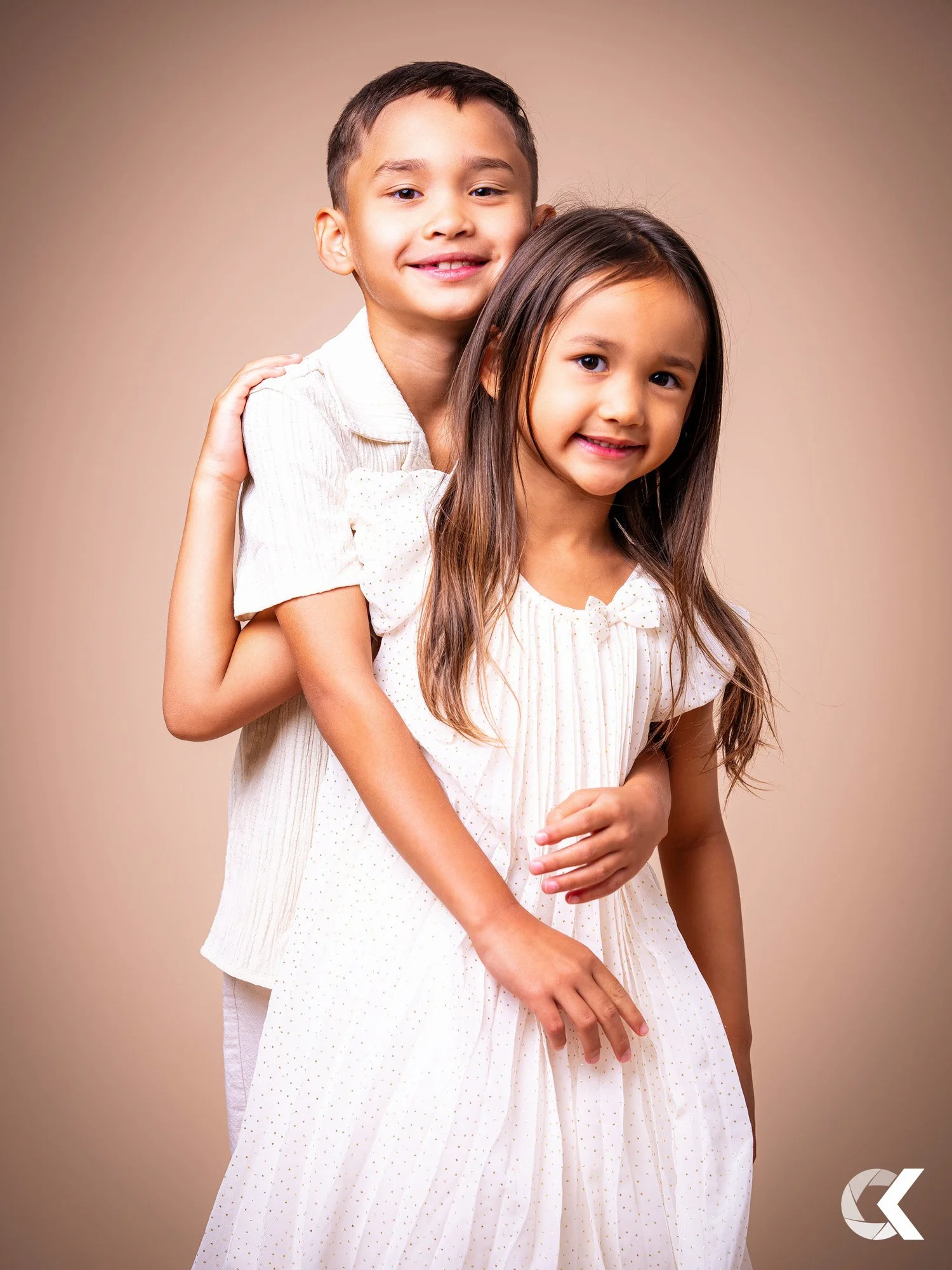 A young boy and girl smiling, with the boy standing behind the girl and embracing her, both wearing light-colored clothes, against a plain beige background.