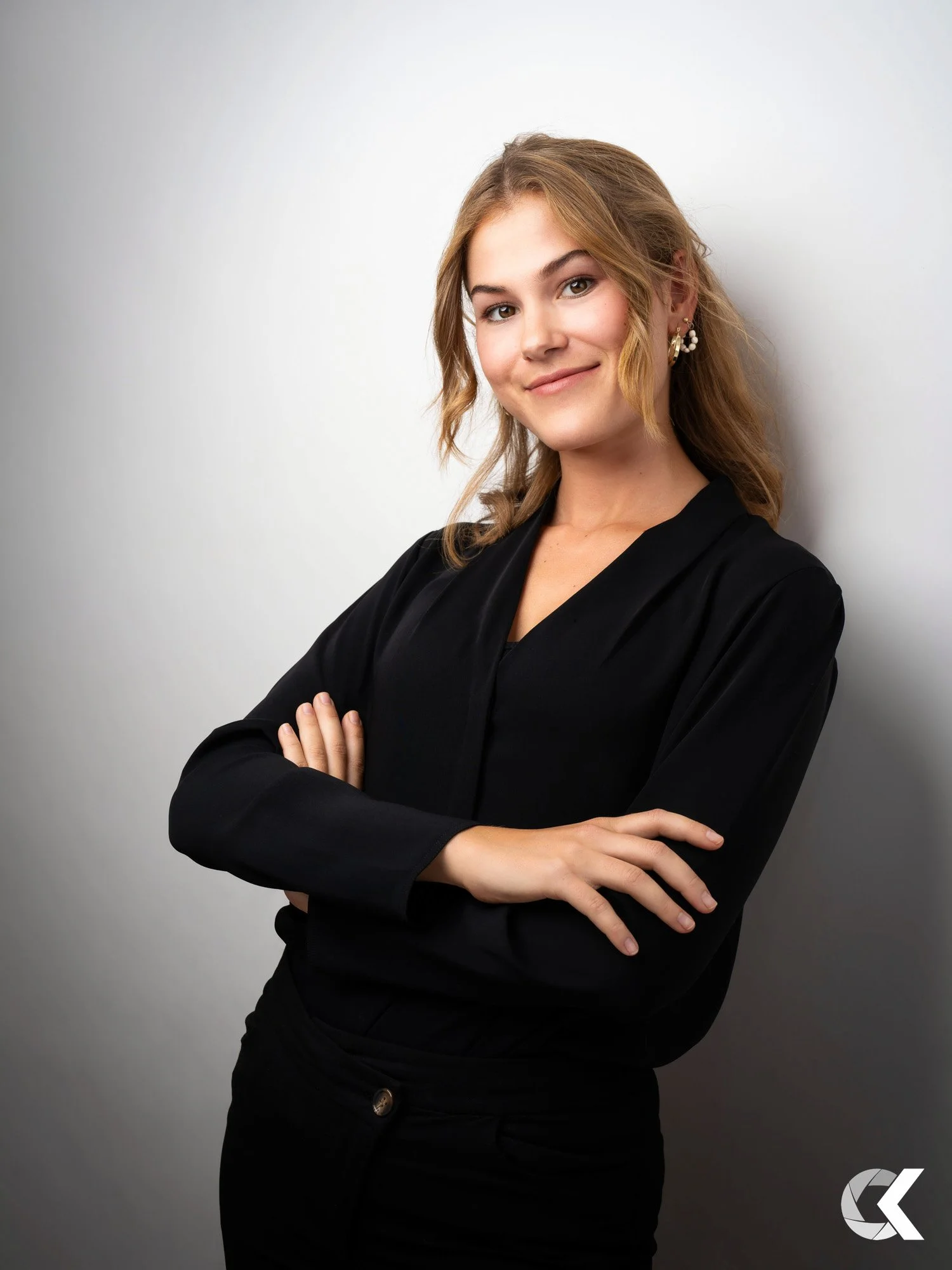 A young woman with wavy blonde hair smiling and crossing her arms, wearing a black long-sleeve top and earrings, standing against a plain white wall.