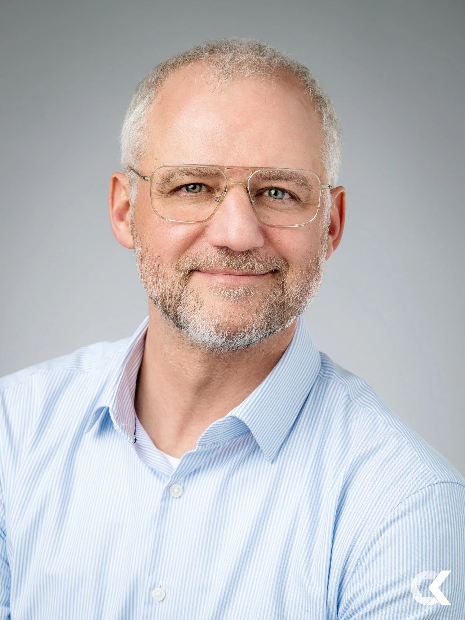 A middle-aged man with gray hair, glasses, and a beard, wearing a light blue striped shirt, smiling slightly against a neutral background.