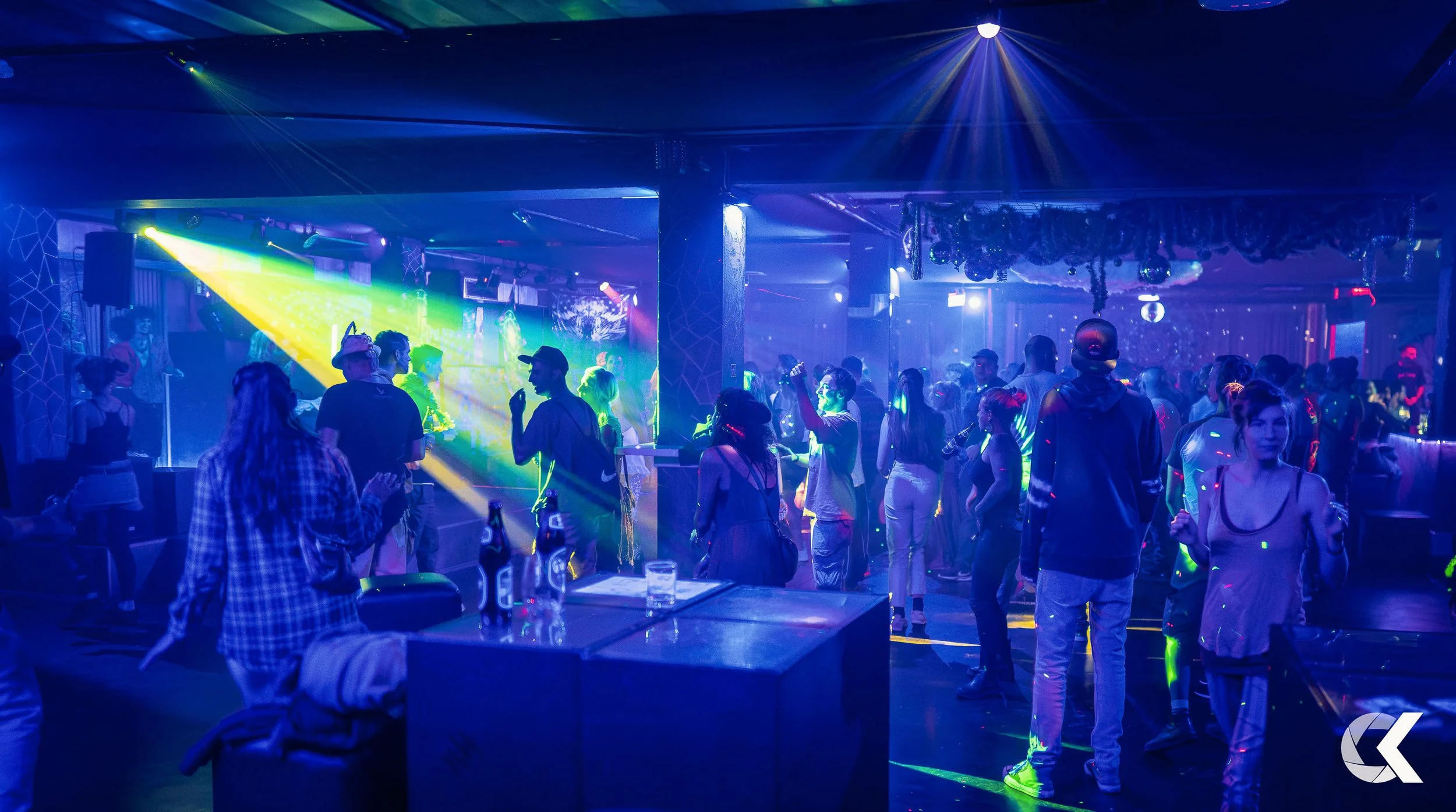 Crowd of people dancing and socializing in a dimly lit nightclub with colorful laser lights and visual effects.