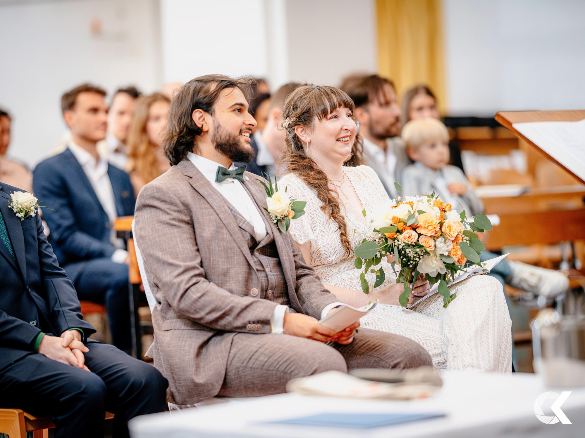A wedding ceremony with a bride and groom seated among guests, smiling and holding programs, with the bride holding a bouquet of flowers.