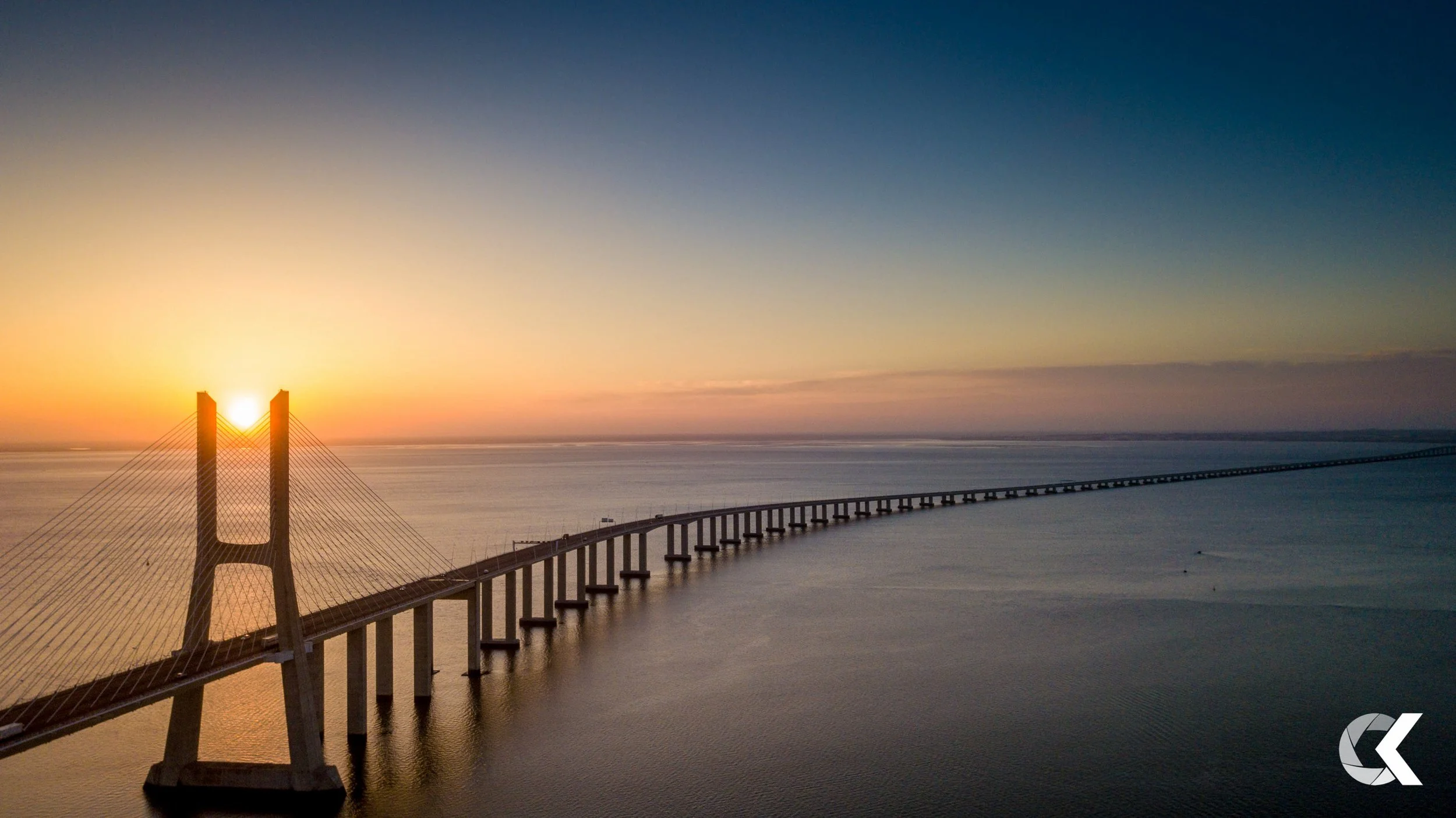 A large suspension bridge extends over water at sunset, with the sun positioned between two tall bridge towers.