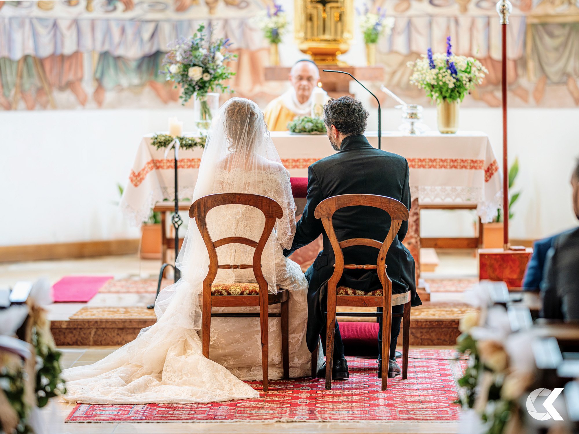 Bride and groom sit in front of altar during wedding ceremony in a church with floral arrangements.