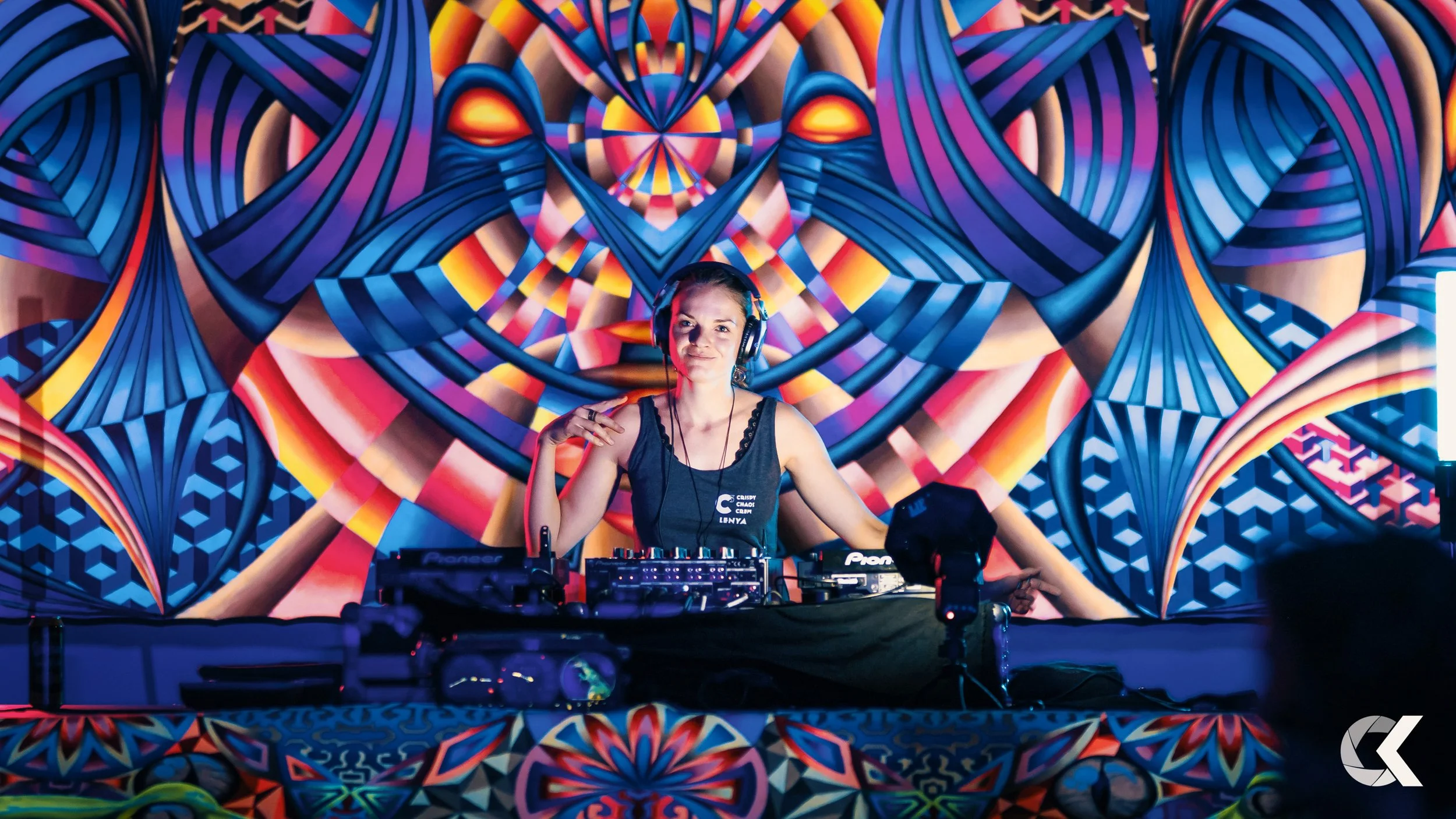 A female DJ stands behind a DJ mixer and turntables on a stage with a vibrant, colorful, geometric, psychedelic background that features symmetrical patterns and abstract shapes.
