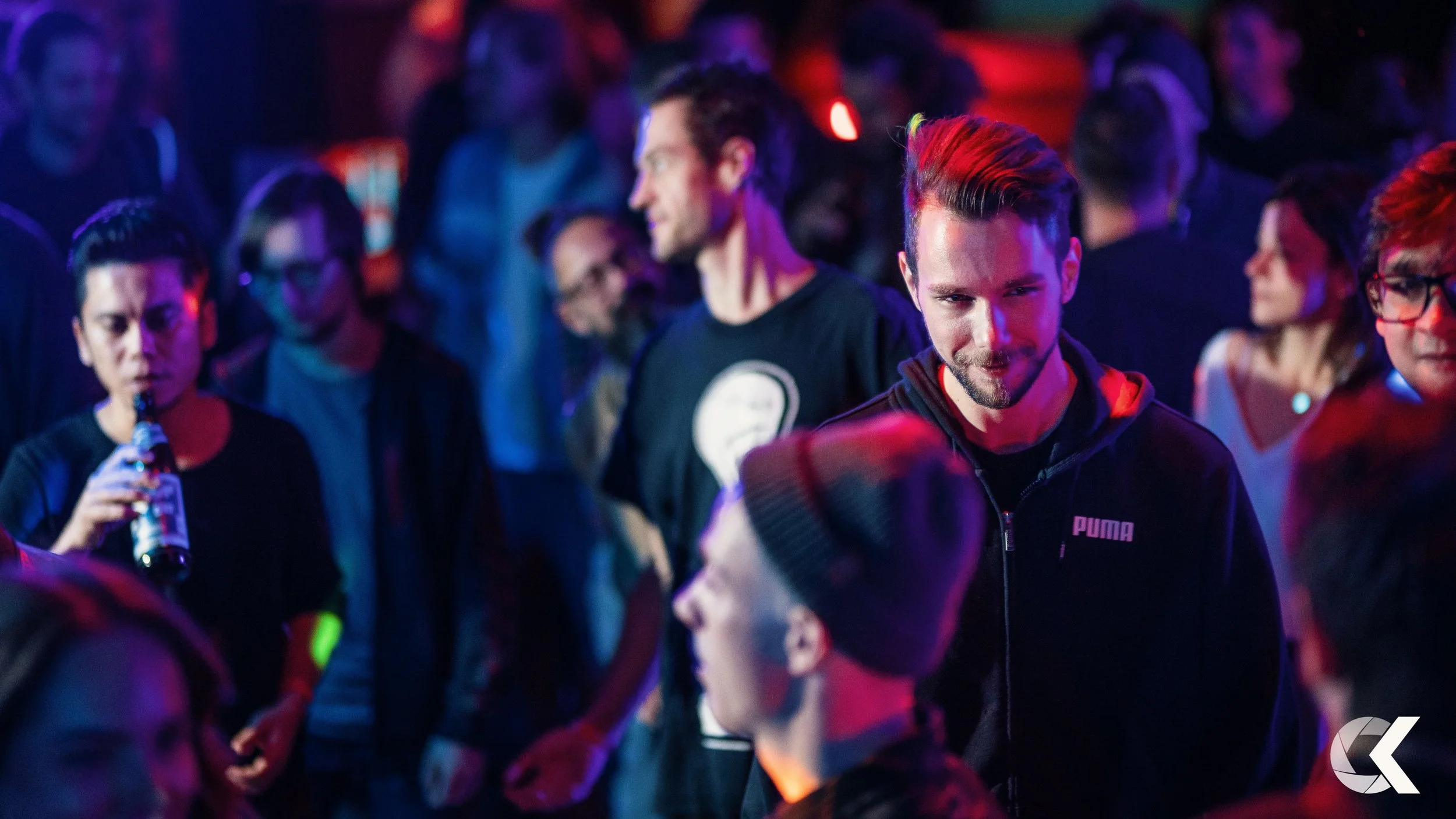 People socializing at a nightclub or party with colorful low lighting, some holding drinks, and engaging in conversations.