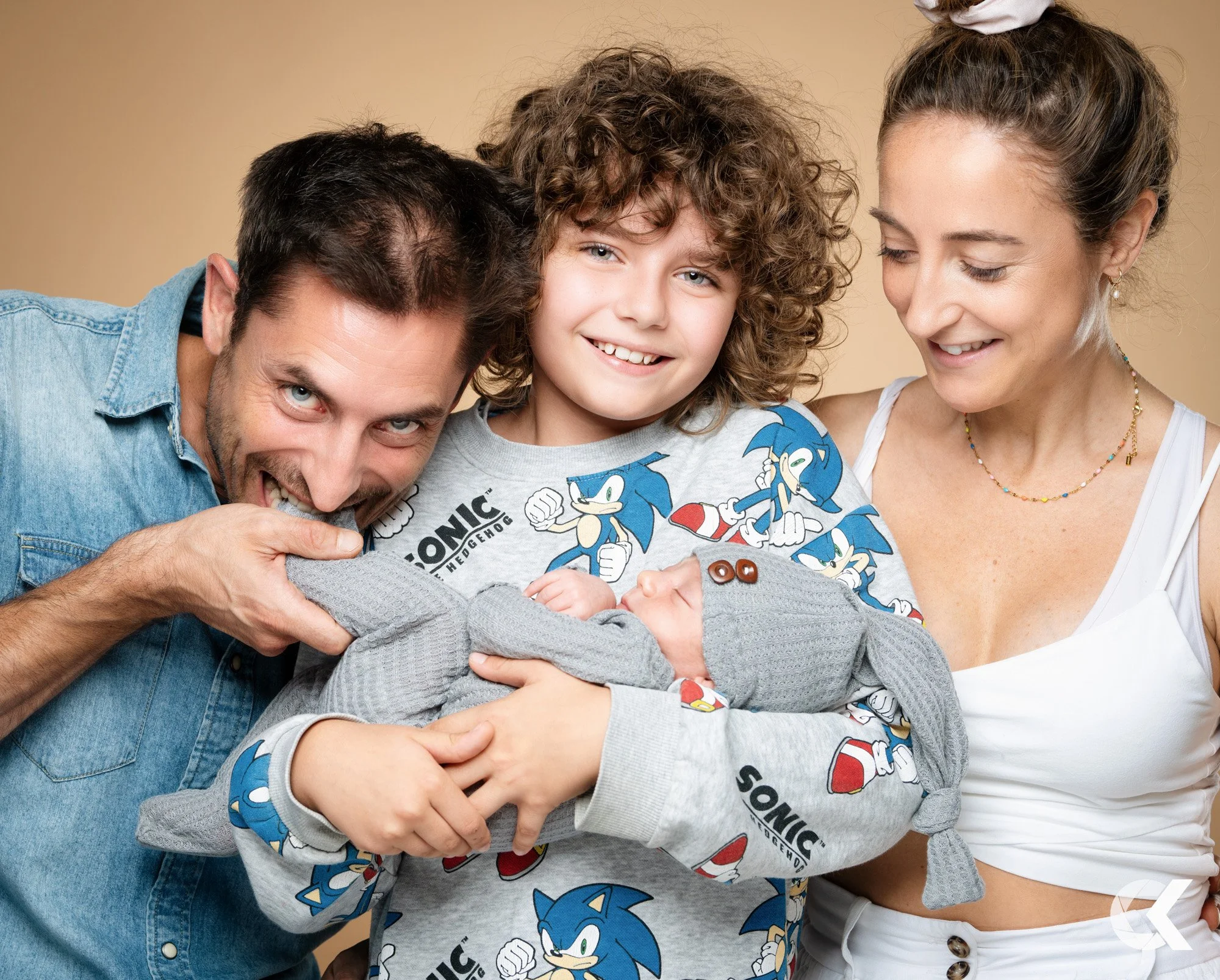 A family of four with light skin celebrating a new baby. The father has dark hair and a beard, and is playfully biting the baby's hand. The mother has her hair tied back and is smiling at the baby. The older child has curly hair and is smiling at the