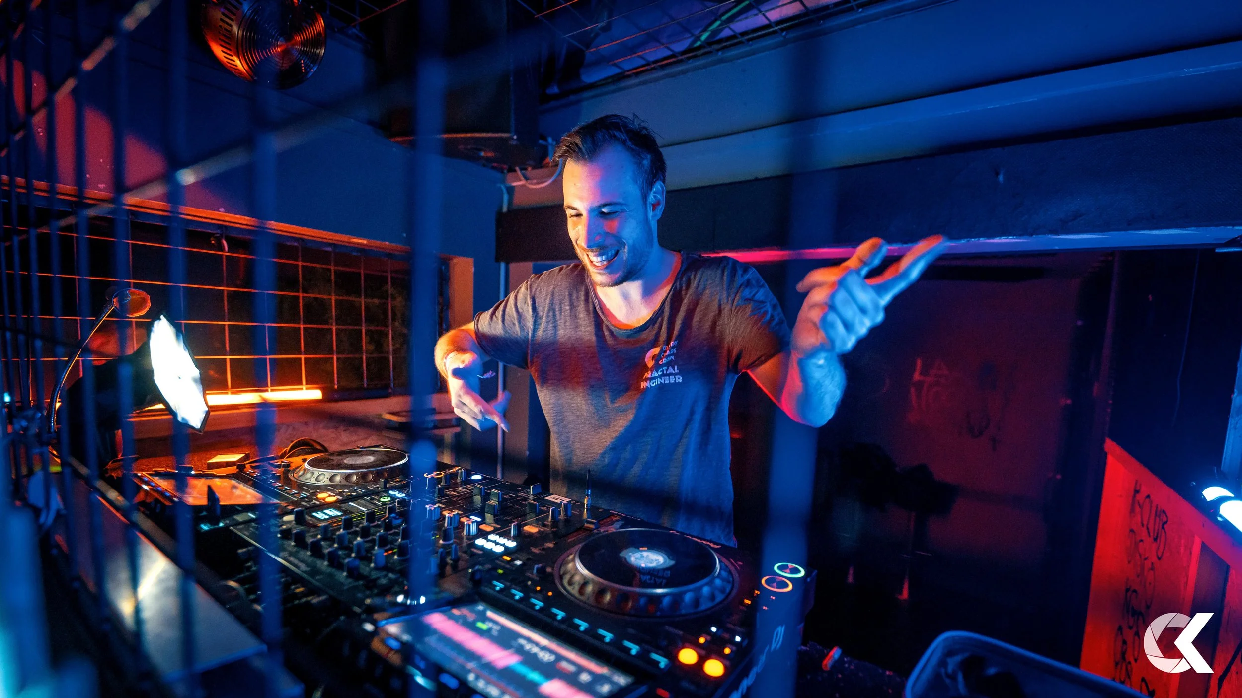 A DJ performing at a nightclub or music venue, smiling and making the peace sign with his right hand, surrounded by DJ equipment illuminated with colorful lights.