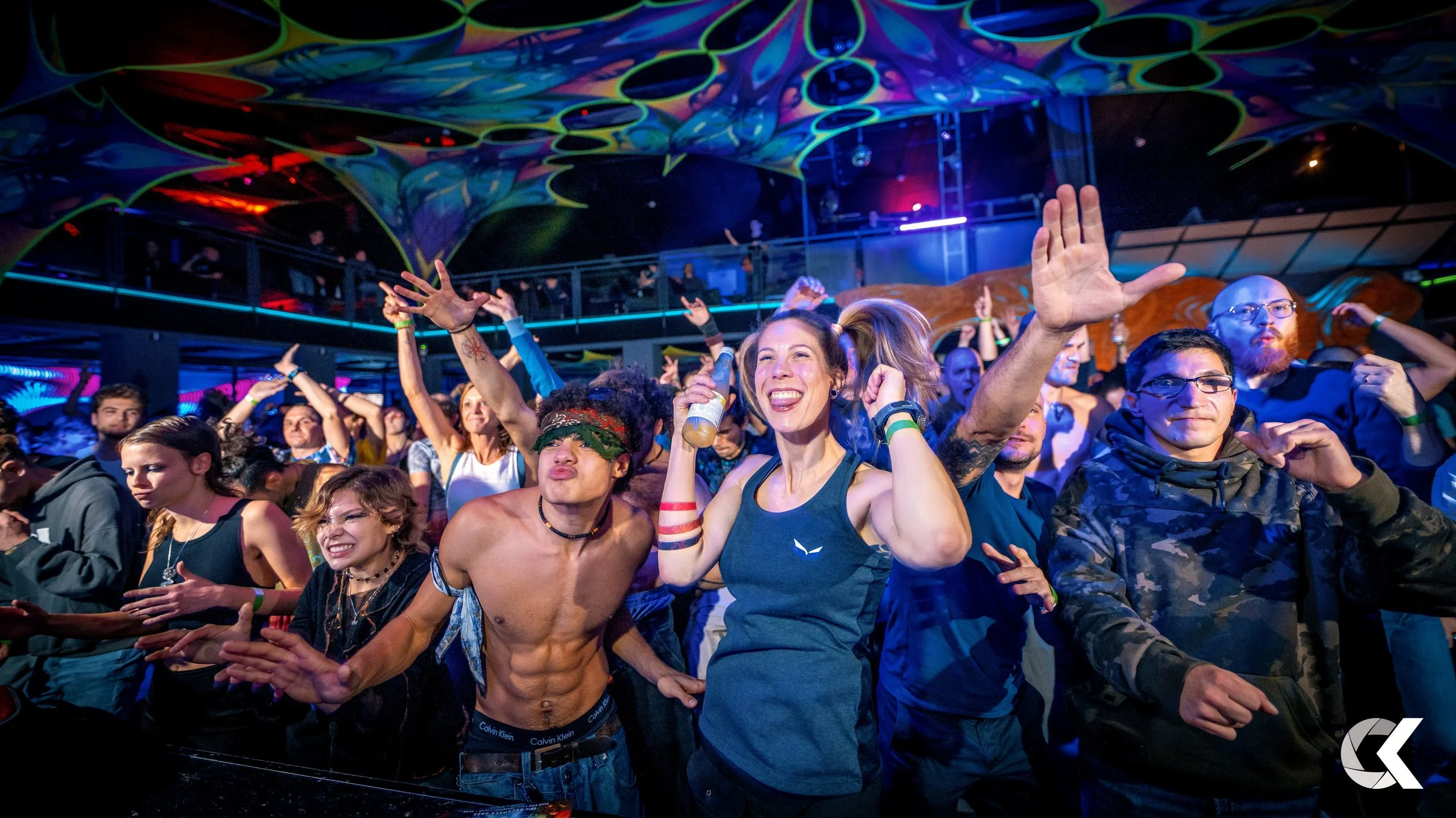 Crowd of people dancing and enjoying a concert at an indoor venue with colorful lighting and a high ceiling.