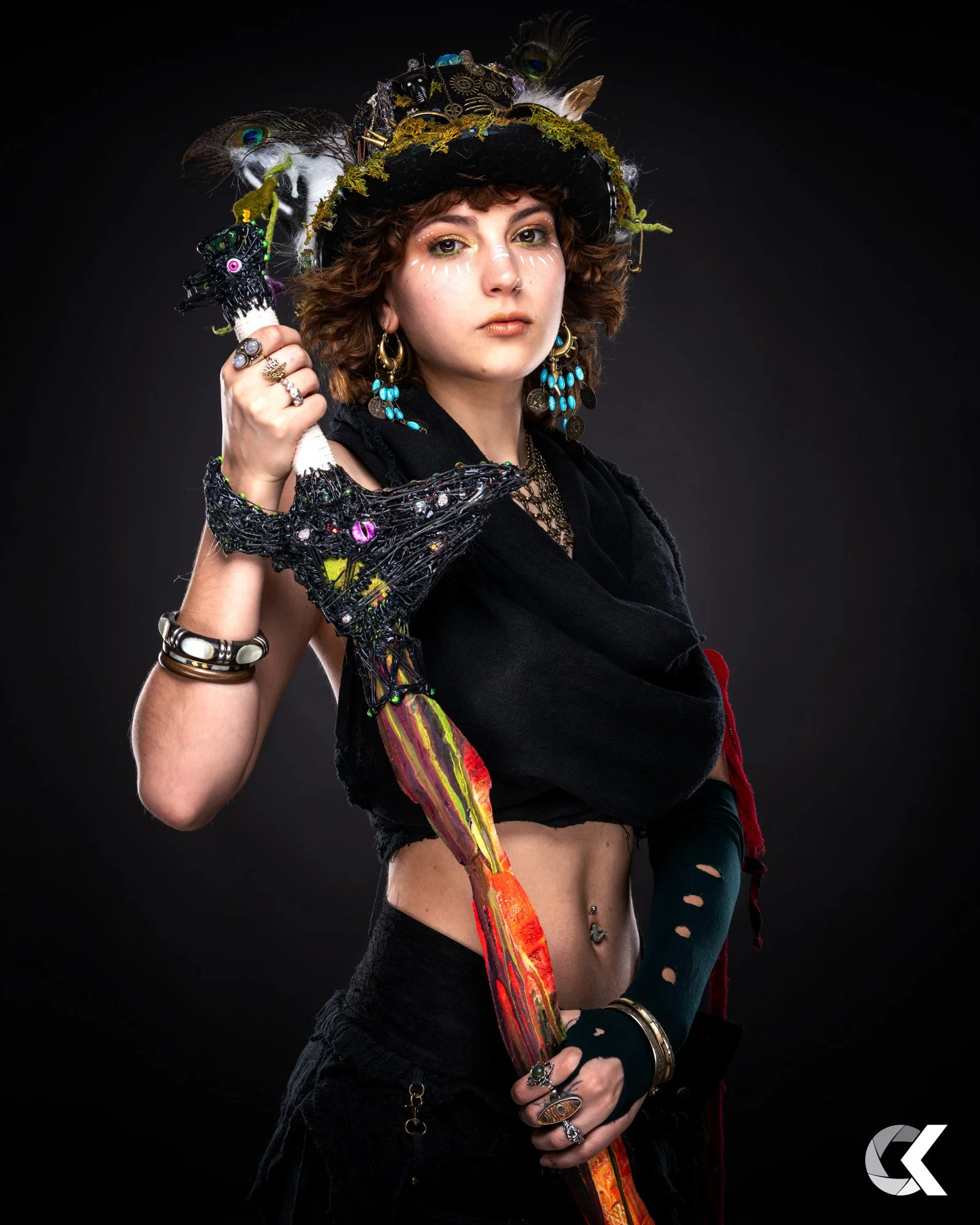 A young woman with curly brown hair wearing a black hat decorated with peacock feathers, jewelry, and face paint, holding a colorful, artistic staff with a dragon head, against a black background.