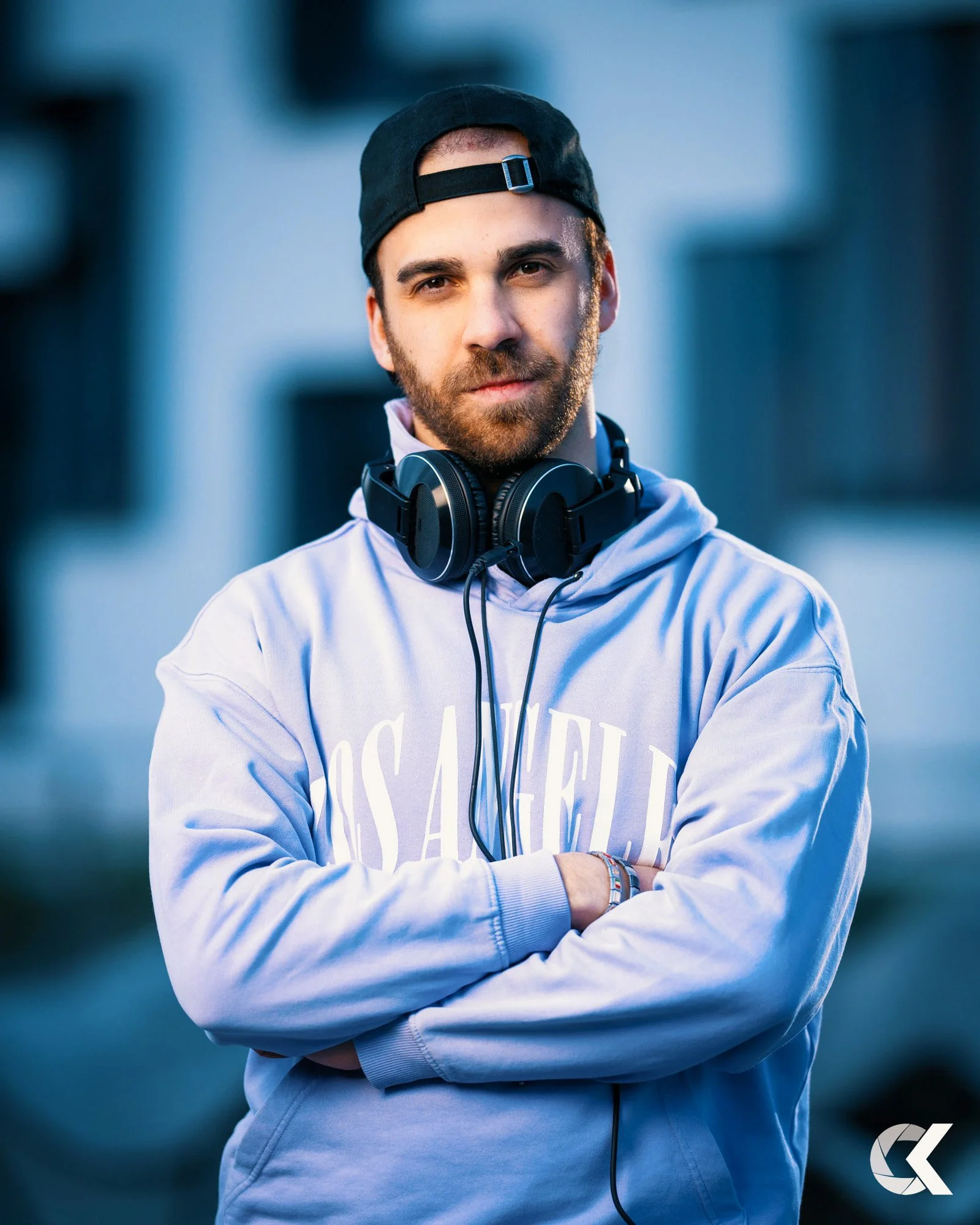 A young man with a beard, wearing a black backwards cap, a light purple Los Angeles hoodie, and headphones around his neck, standing outdoors with arms crossed and a serious expression.