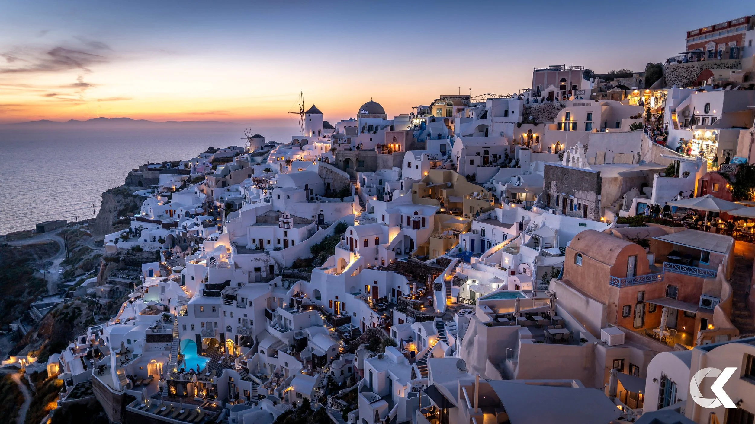 Sunset over the white buildings of a Greek island village, with the sea in the background and string lights illuminating the streets and terraces.