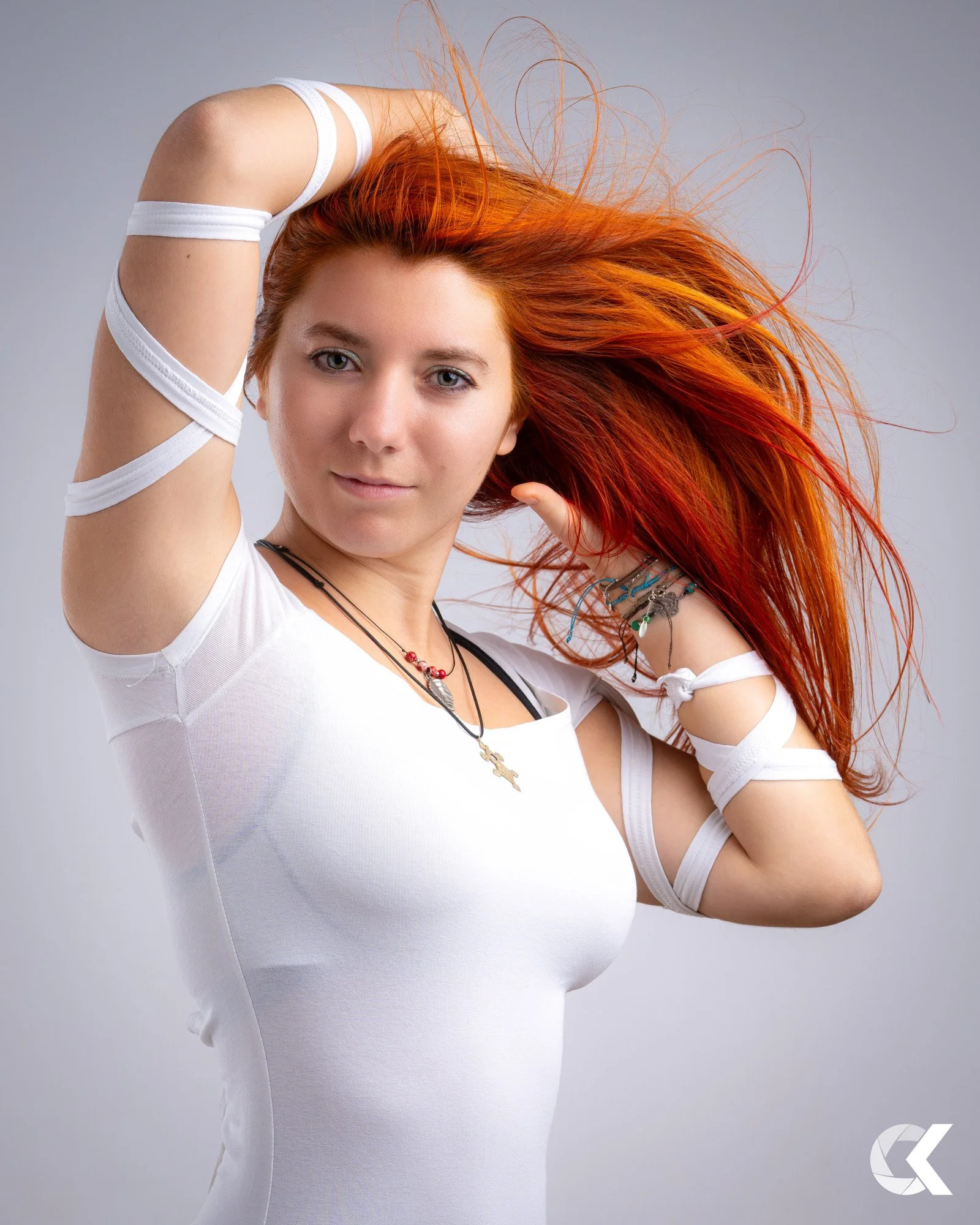 Portrait einer jungen Frau mit langen, roten Haaren, die sich mit einer Hand im Haar und der anderen am Nacken fasst, trägt ein weißes T-Shirt und mehrere Halsketten, auf einem grauen Hintergrund.