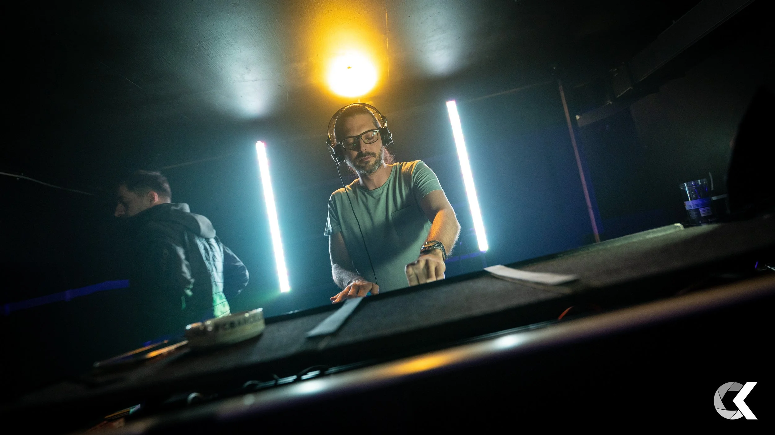 DJ with headphones and glasses performing in a dark club with neon lights.