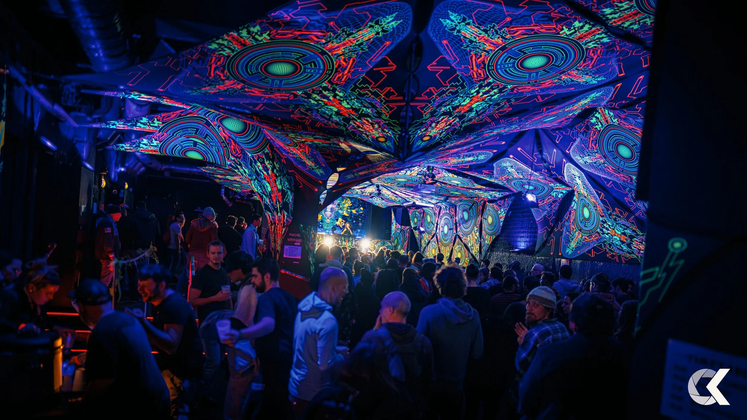 A crowded indoor concert or event with vibrant, glowing, psychedelic light projections on the ceiling and walls. People are standing and watching a performance on stage, which is illuminated with bright lights.