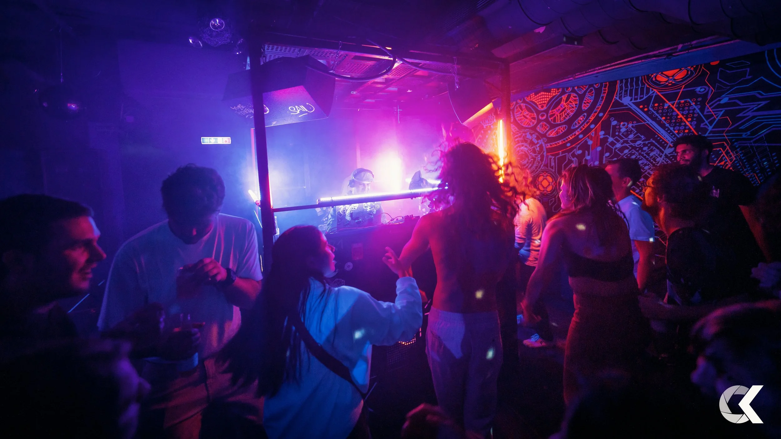 People dancing and socializing in a dark, colorful nightclub with neon purple and orange lights and psychedelic wall art.
