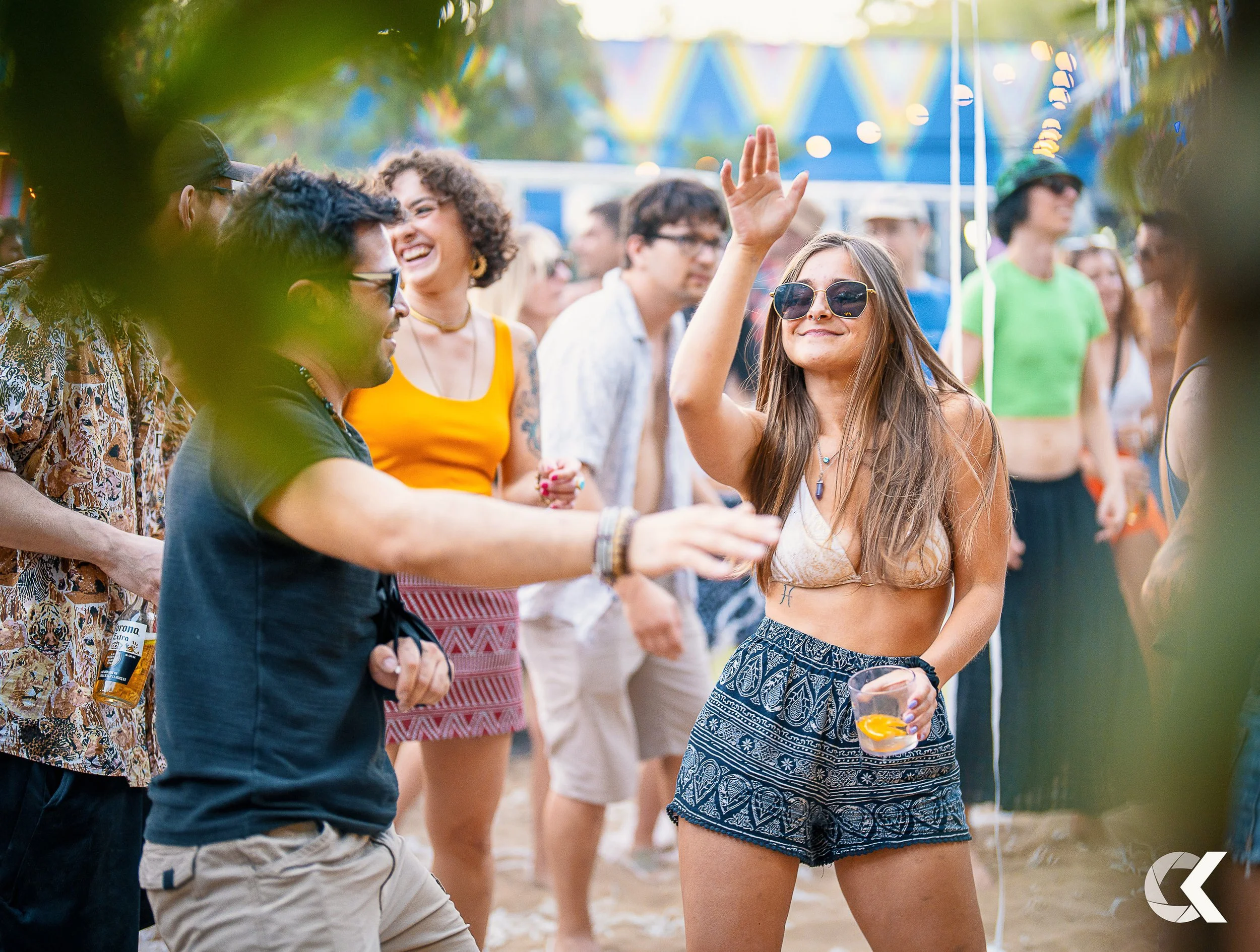 People dancing and enjoying themselves at an outdoor music festival or party, with a woman in sunglasses smiling and raising her hand in the center, holding a drink in her other hand.